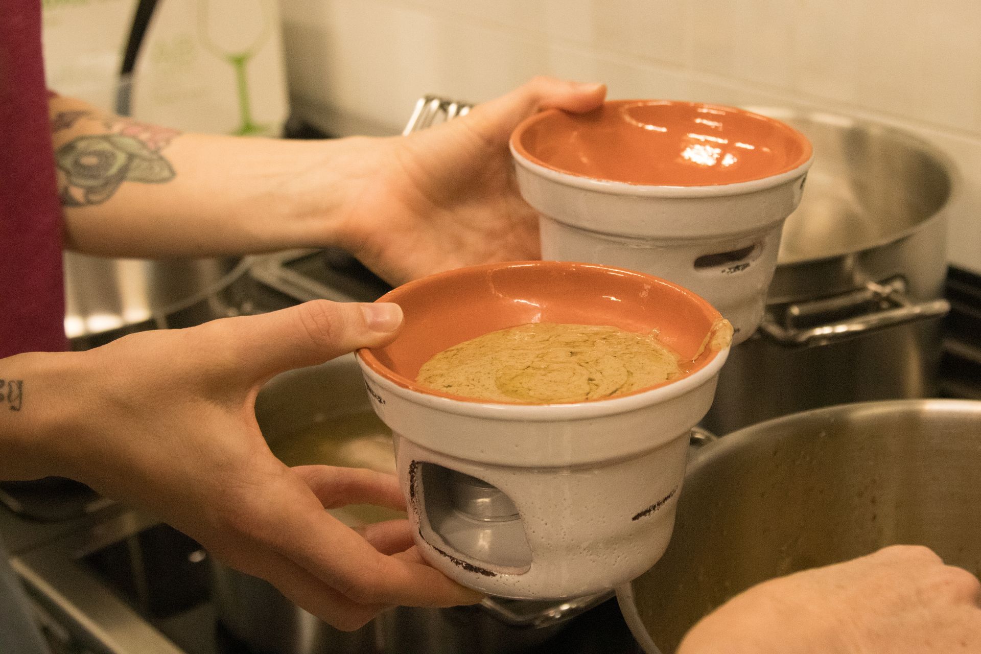 Due mani sorreggono pentole per fonduta in terracotta con fondo in ceramica sopra i fornelli della cucina.