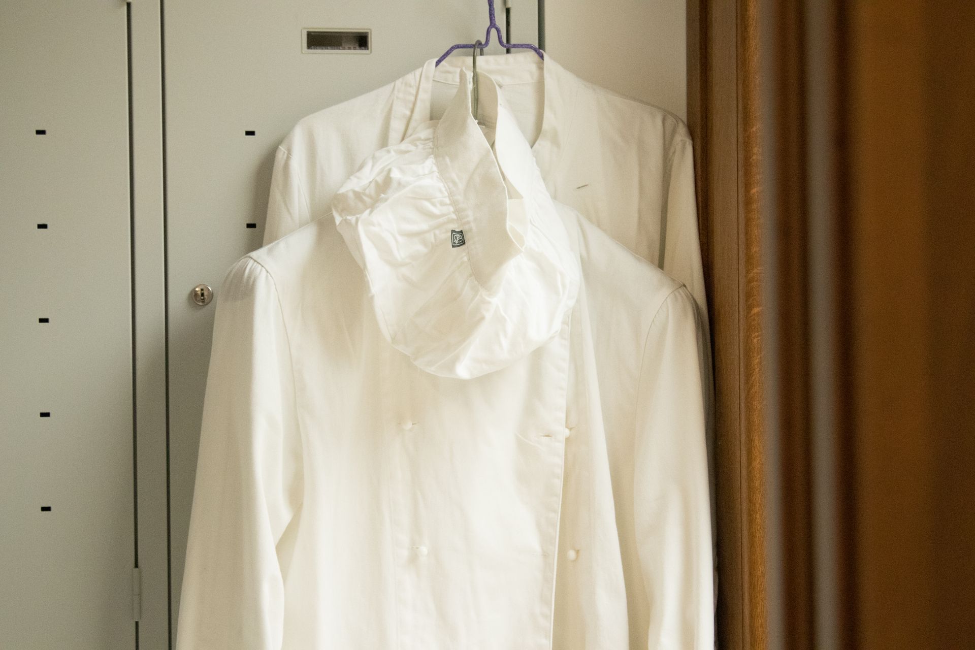 Due camici da laboratorio bianchi con cappuccio sono appesi a una gruccia di metallo contro la porta di un armadietto grigio.