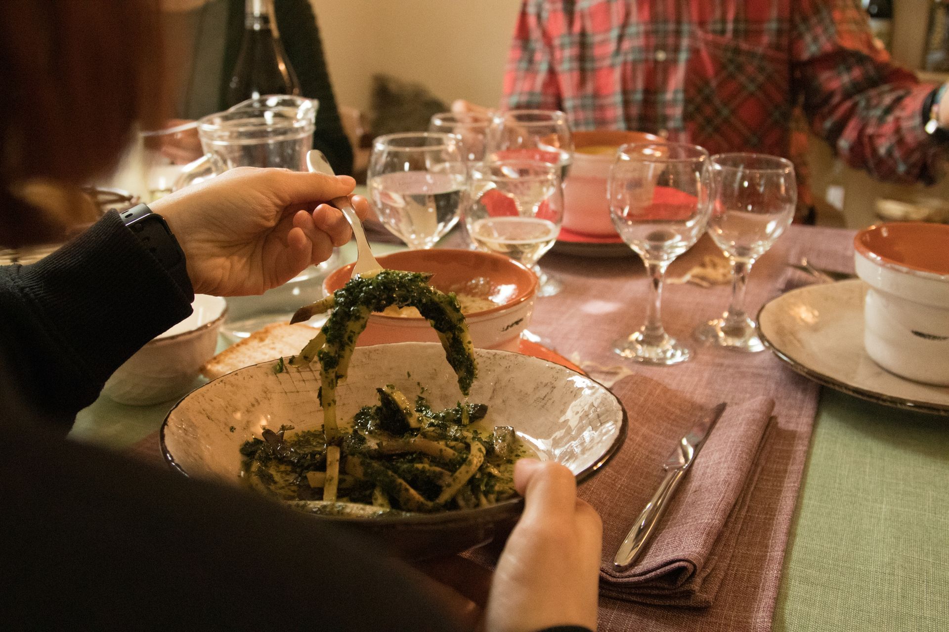 Una persona prende una forchettata di verdure verdi da una ciotola su un tavolo apparecchiato con bicchieri da vino e tovaglioli decorati.