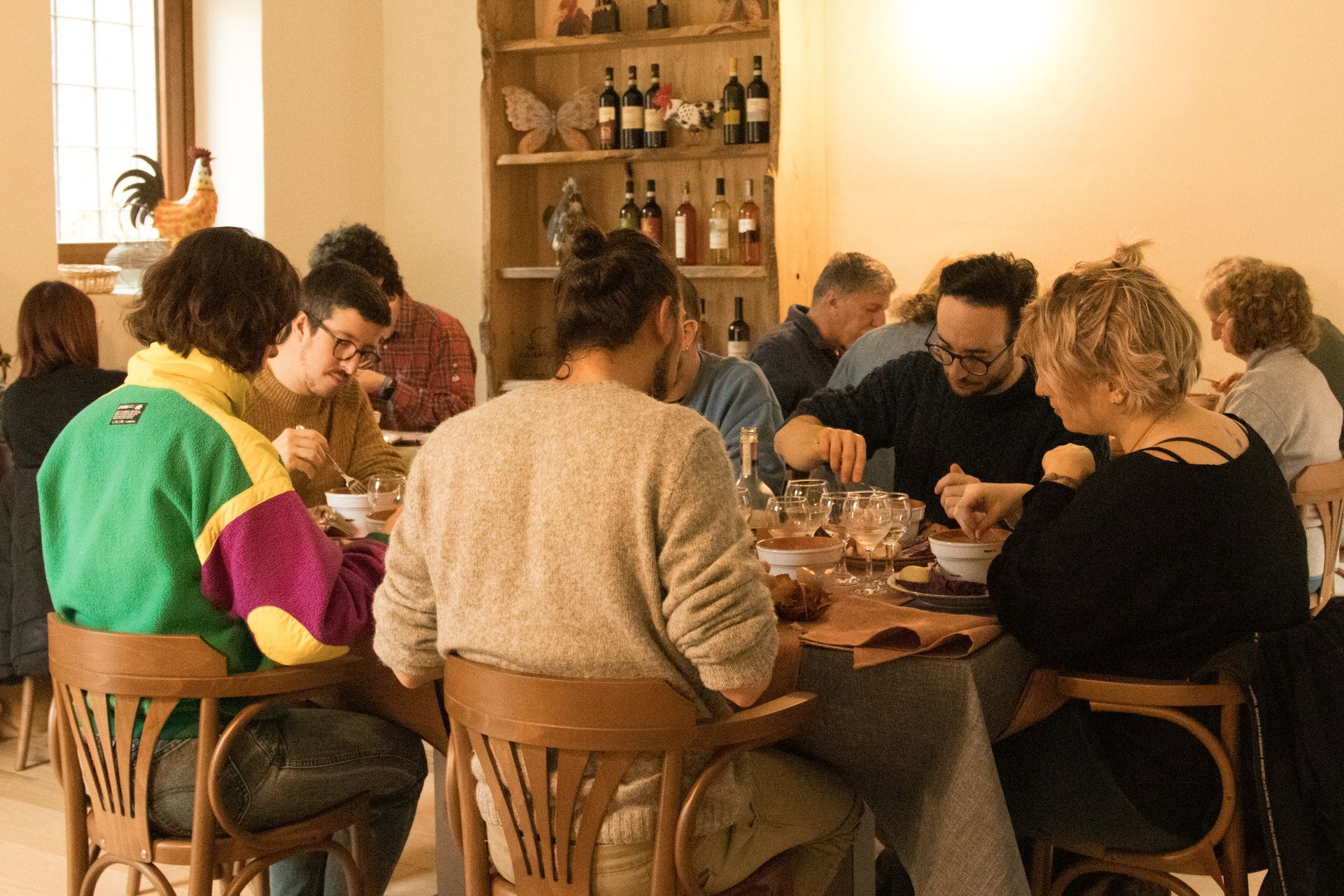 Un gruppo eterogeneo di persone sedute attorno a un tavolo in una sala da pranzo rustica, condividono un pasto e una conversazione.