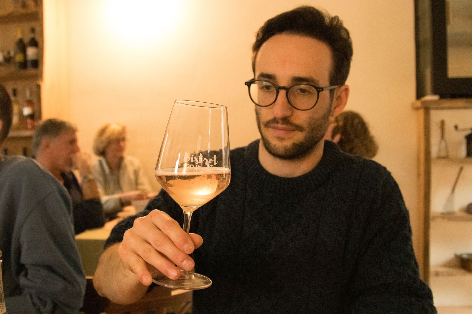 Una persona con un maglione scuro tiene in mano un bicchiere di vino e lo osserva in un ristorante.