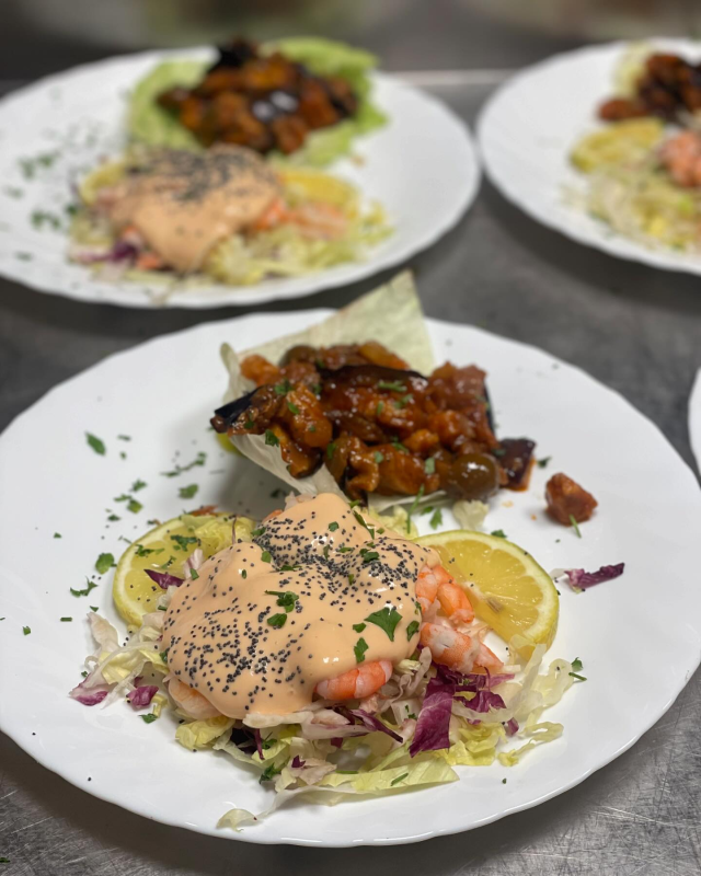 Su un tavolo è impiattata un'insalata di gamberi con fette di limone e verdure saltate, con altri piatti sullo sfondo.