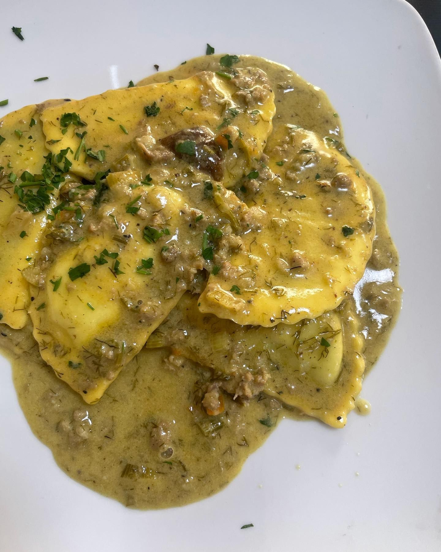 Ravioli ricoperti di formaggio in salsa verde cremosa, serviti su un piatto bianco e guarniti con erbe aromatiche tritate.