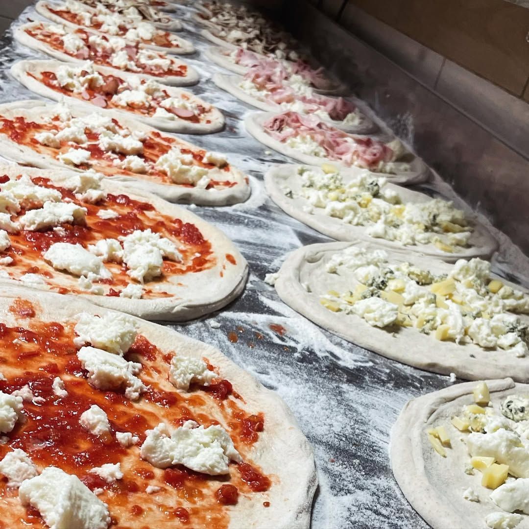 Vassoi di pizze crude condite con salsa di pomodoro e formaggio su una superficie di lavoro infarinata.