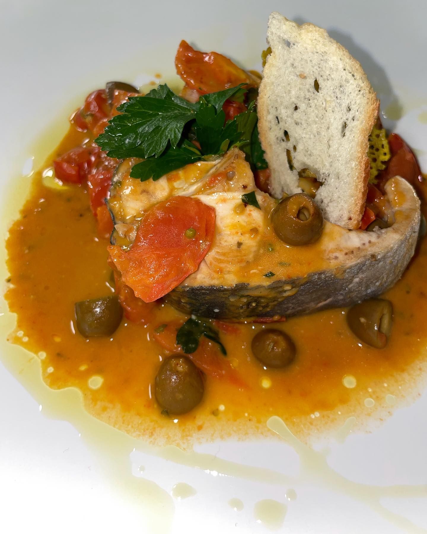 Spezzatino di pomodoro e pesce con olive e prezzemolo, servito su un piatto bianco con una fetta di pane.