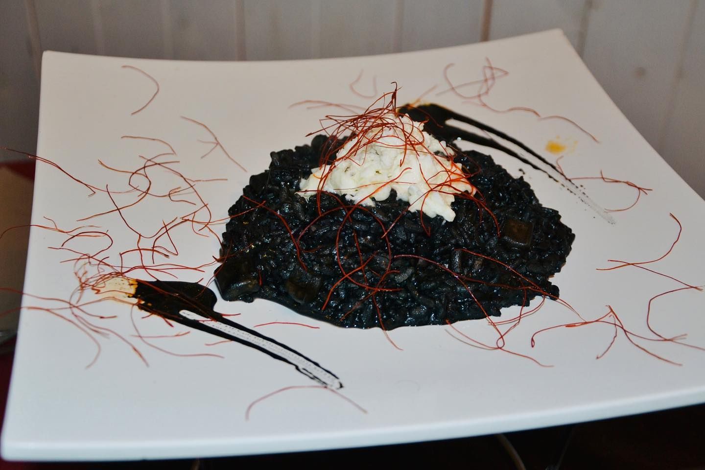 Un mucchietto di riso nero su un piatto bianco, guarnito con una decorazione bianca e una salsa rossa che lo avvolge.