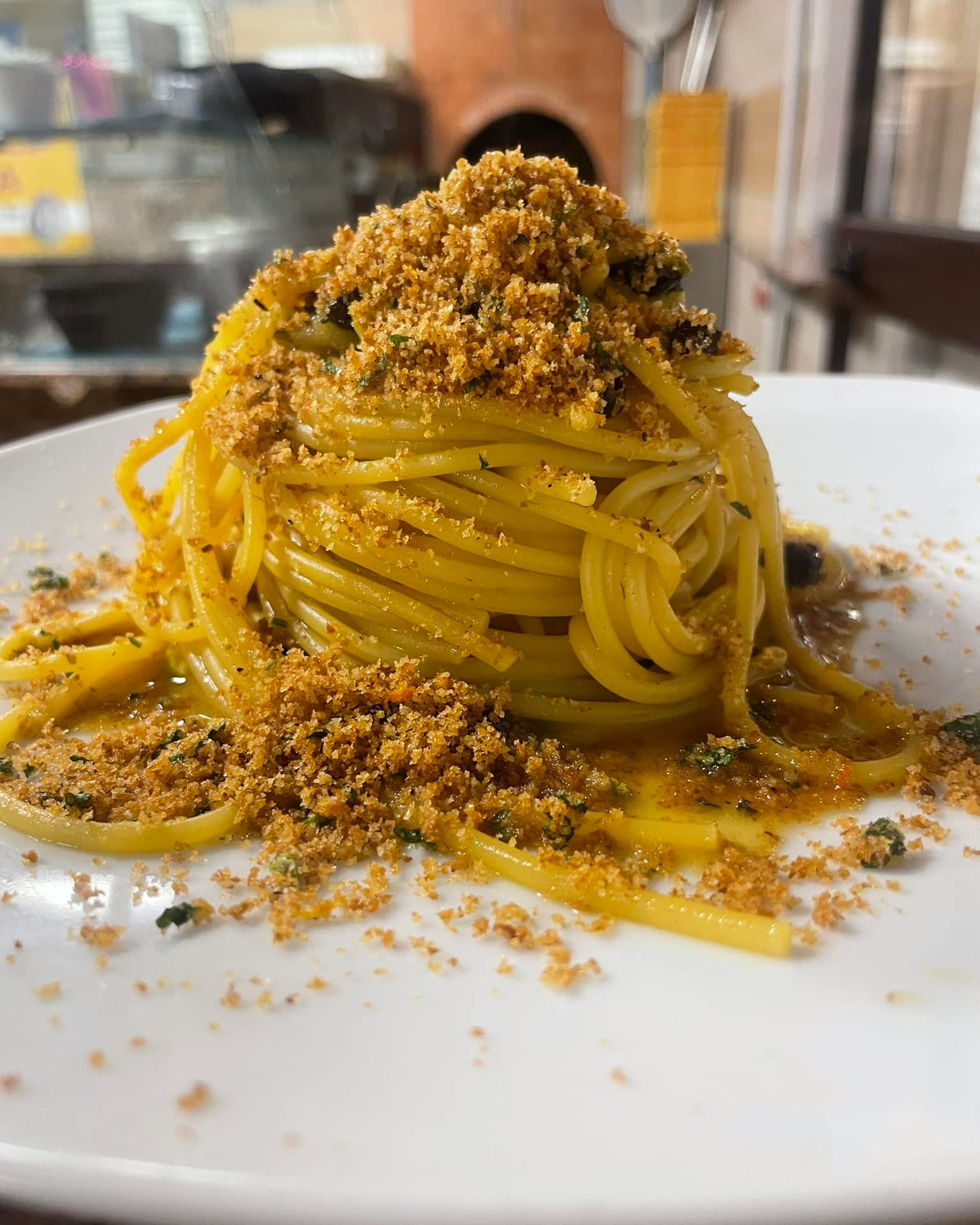 Spaghetti conditi con pangrattato su un piatto bianco in un ambiente da ristorante.