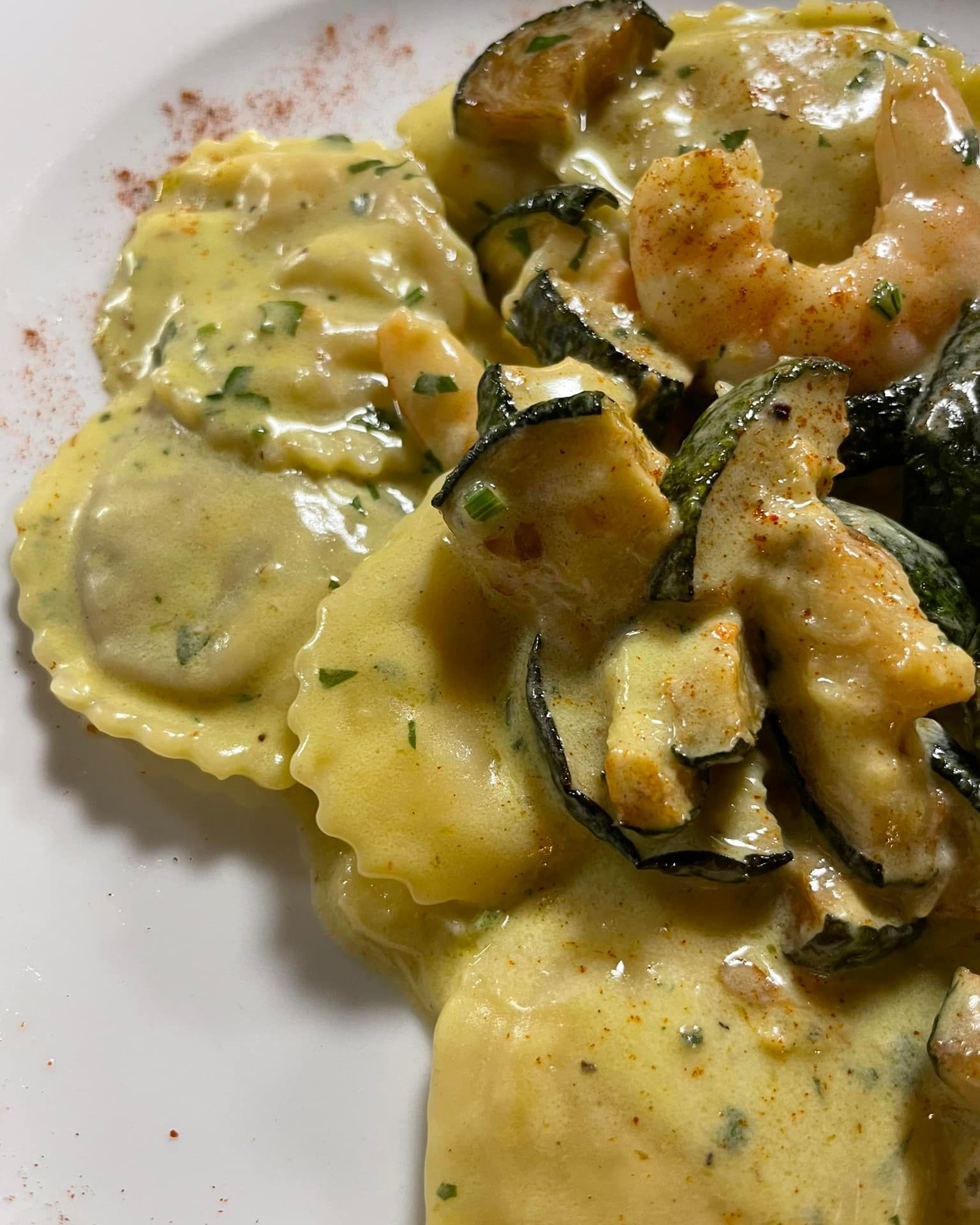 Ravioli cremosi conditi con gamberi e zucchine in salsa gialla alle erbe aromatiche, serviti su un piatto bianco.