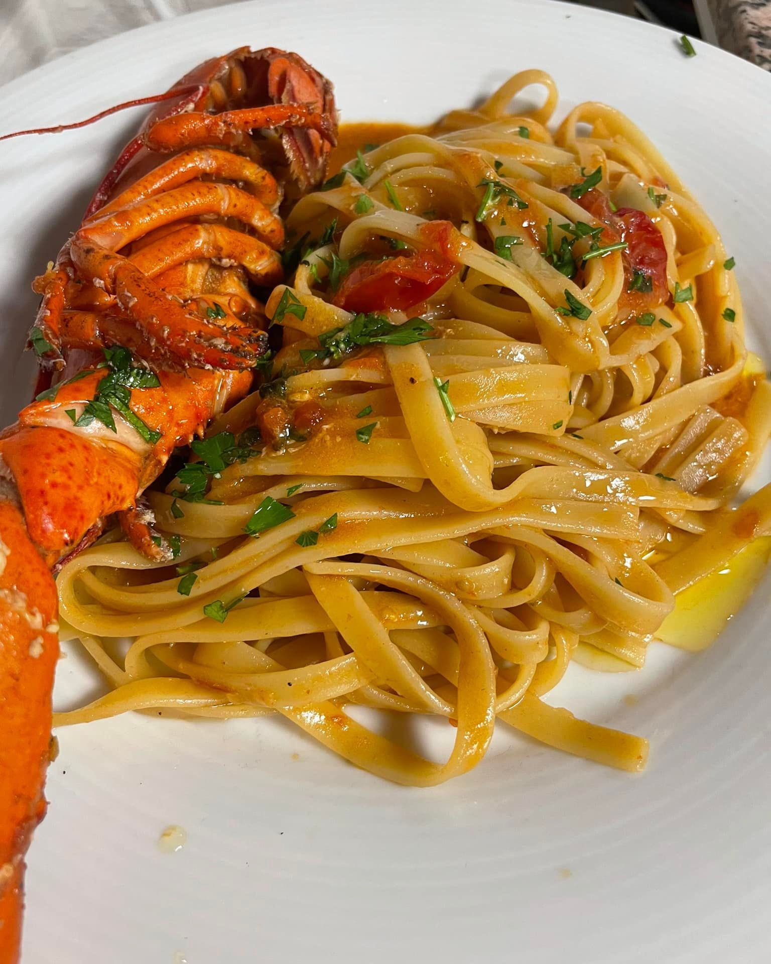 Coda di aragosta con linguine in una ciotola bianca, guarnita con erbe aromatiche e pezzetti di pomodoro.