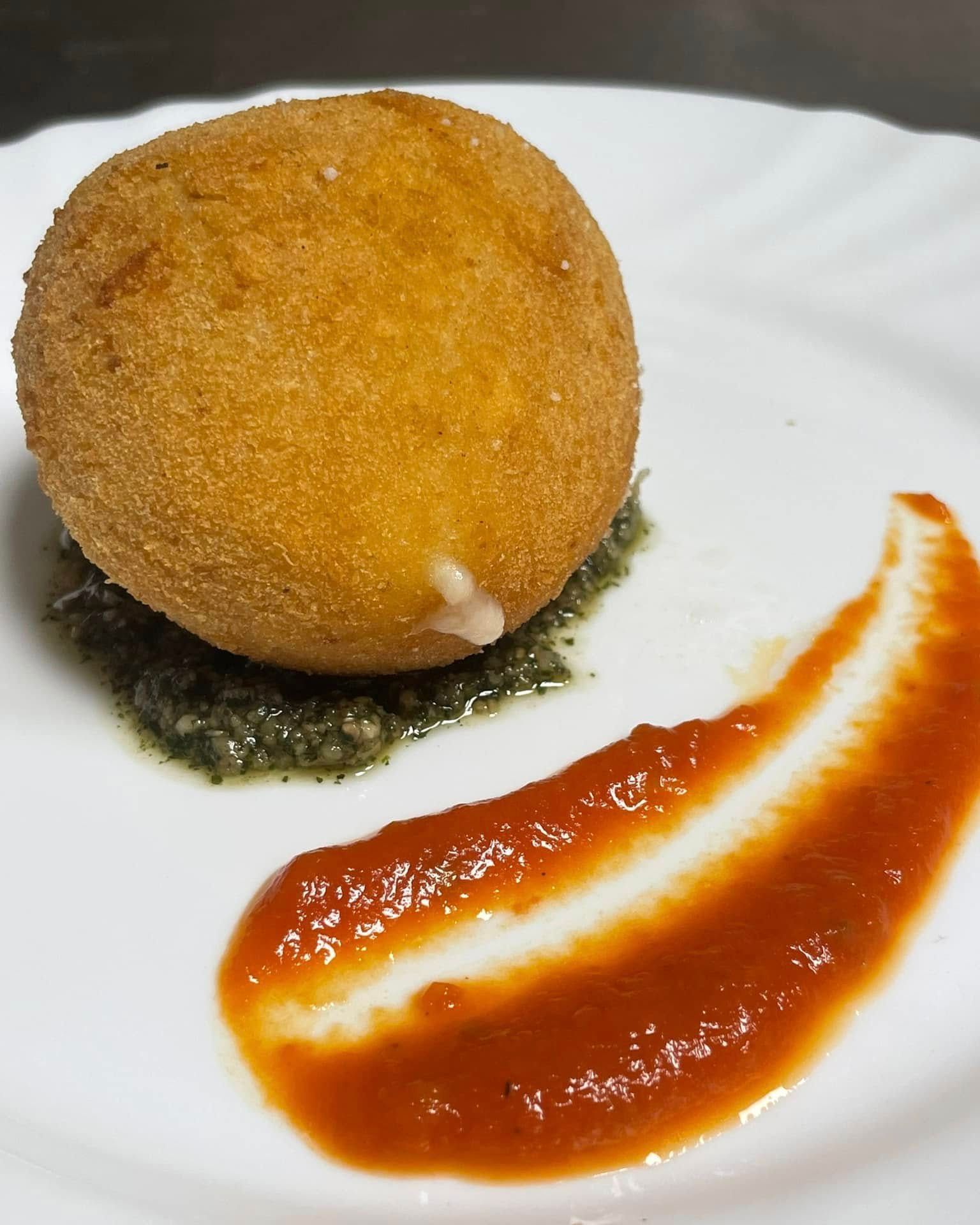 Pallina fritta dorata su un piatto bianco con salsa verde e una spruzzata di salsa rossa.
