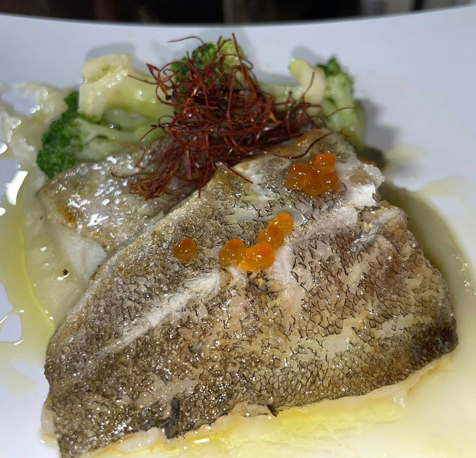 Filetto di pesce servito con broccoli, salsa cremosa e guarnizione d'arancia su un piatto bianco.