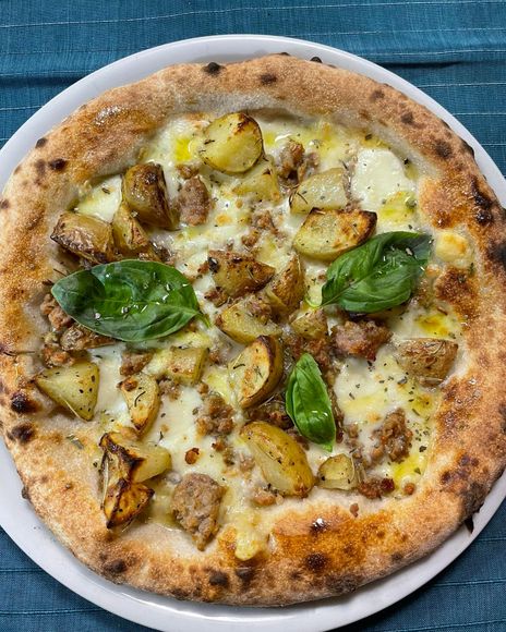 Pizza cotta nel forno a legna condita con patate, basilico e formaggio fuso, servita su un piatto.