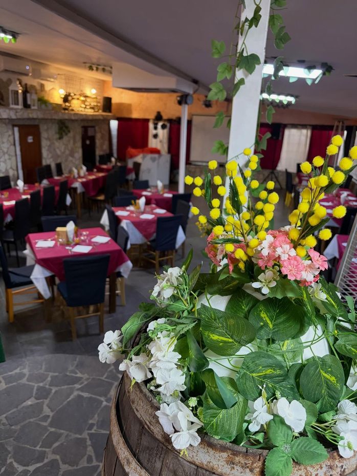 Sala da pranzo rustica di un ristorante con tovaglie rosse e una grande composizione floreale in una botte in primo piano.