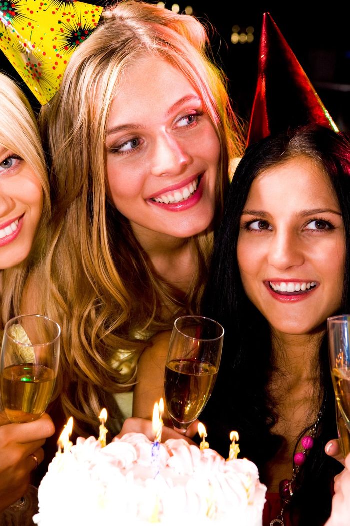 Tre donne sorridenti con calici di champagne accanto a una torta di compleanno e cappellini da festa.