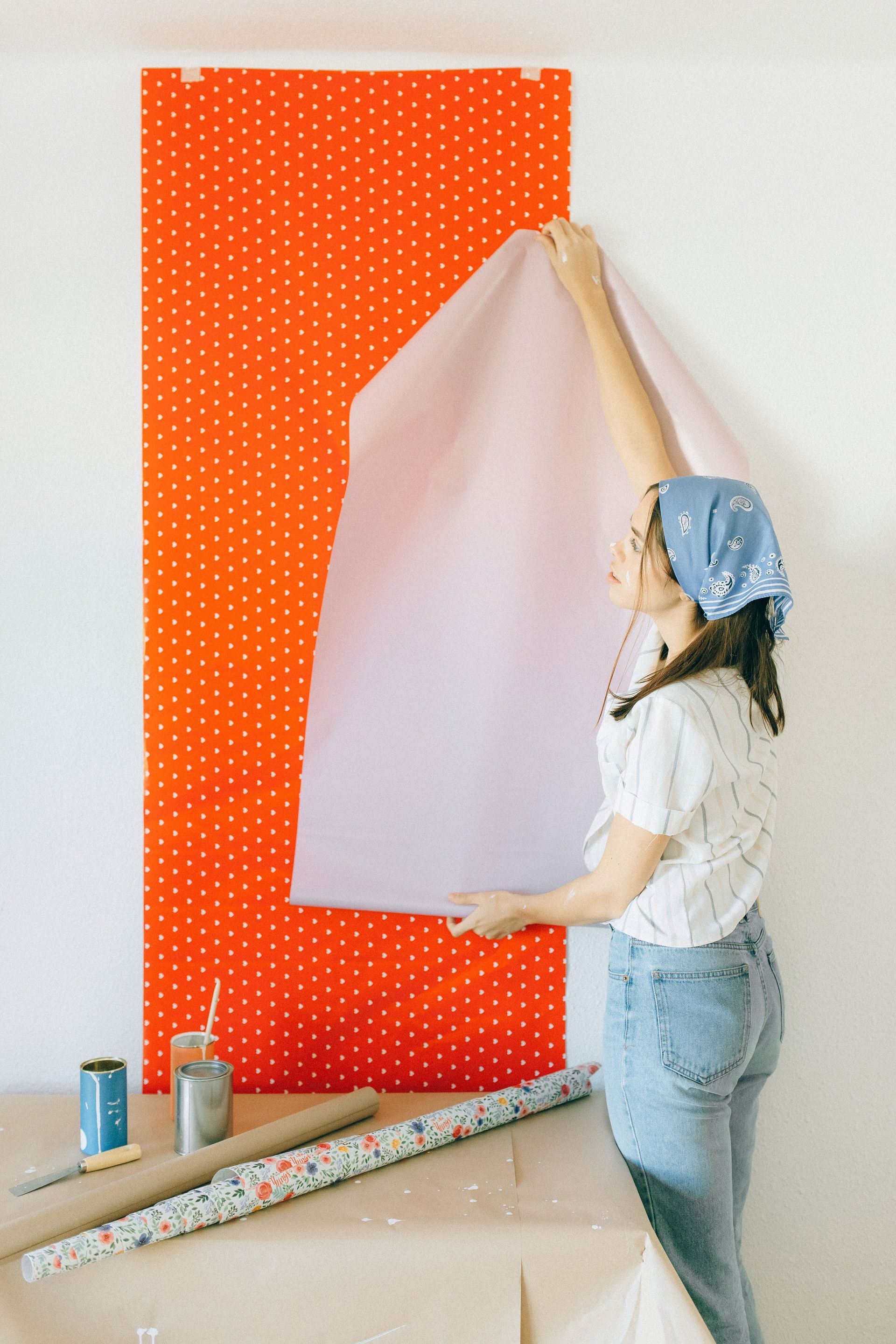 Woman hanging white wallpaper panel over orange polka-dot wallpaper in a room renovation