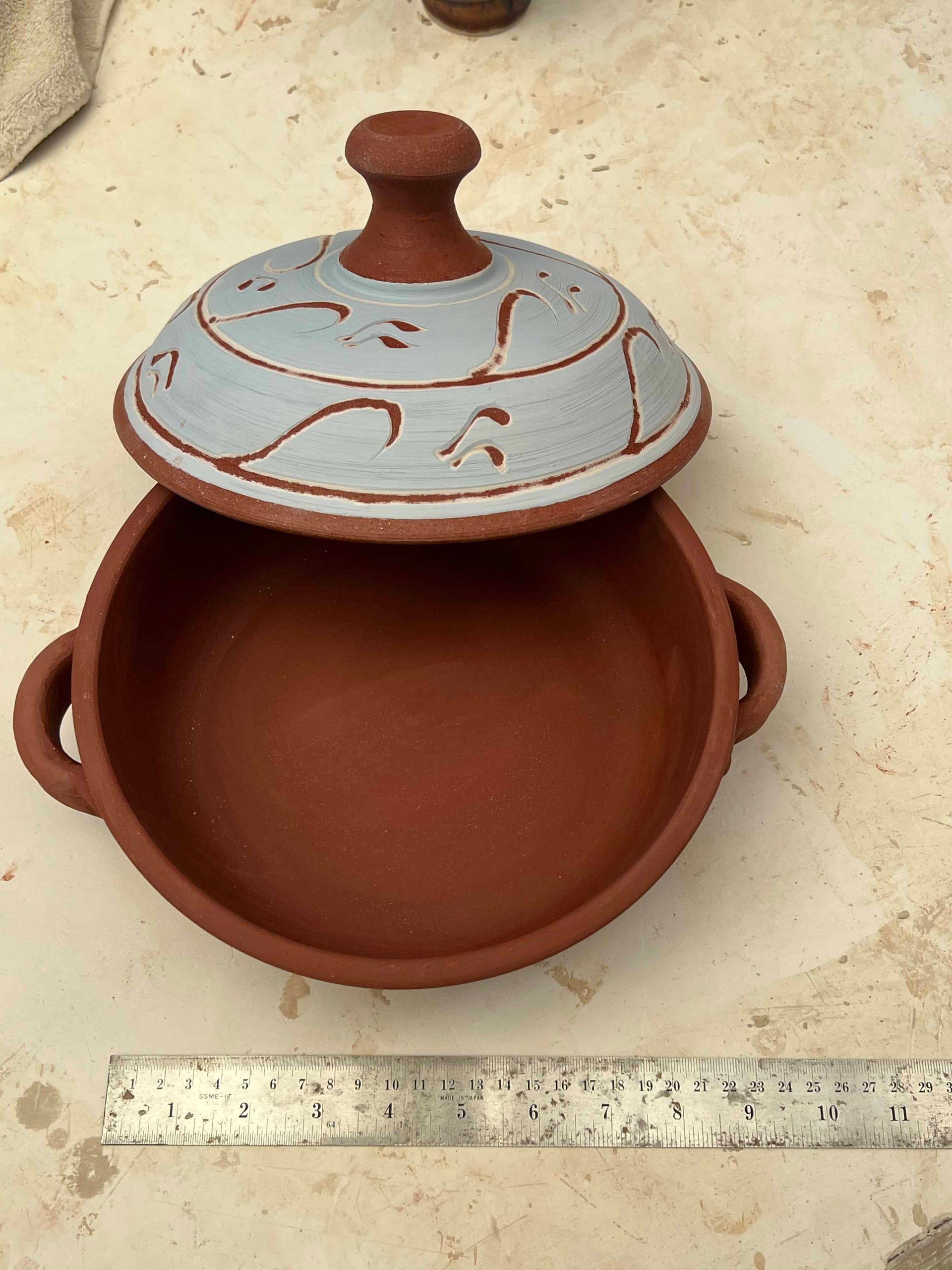 Terracotta pot with a light blue lid that has intricate designs on it, sitting on a white countertop 