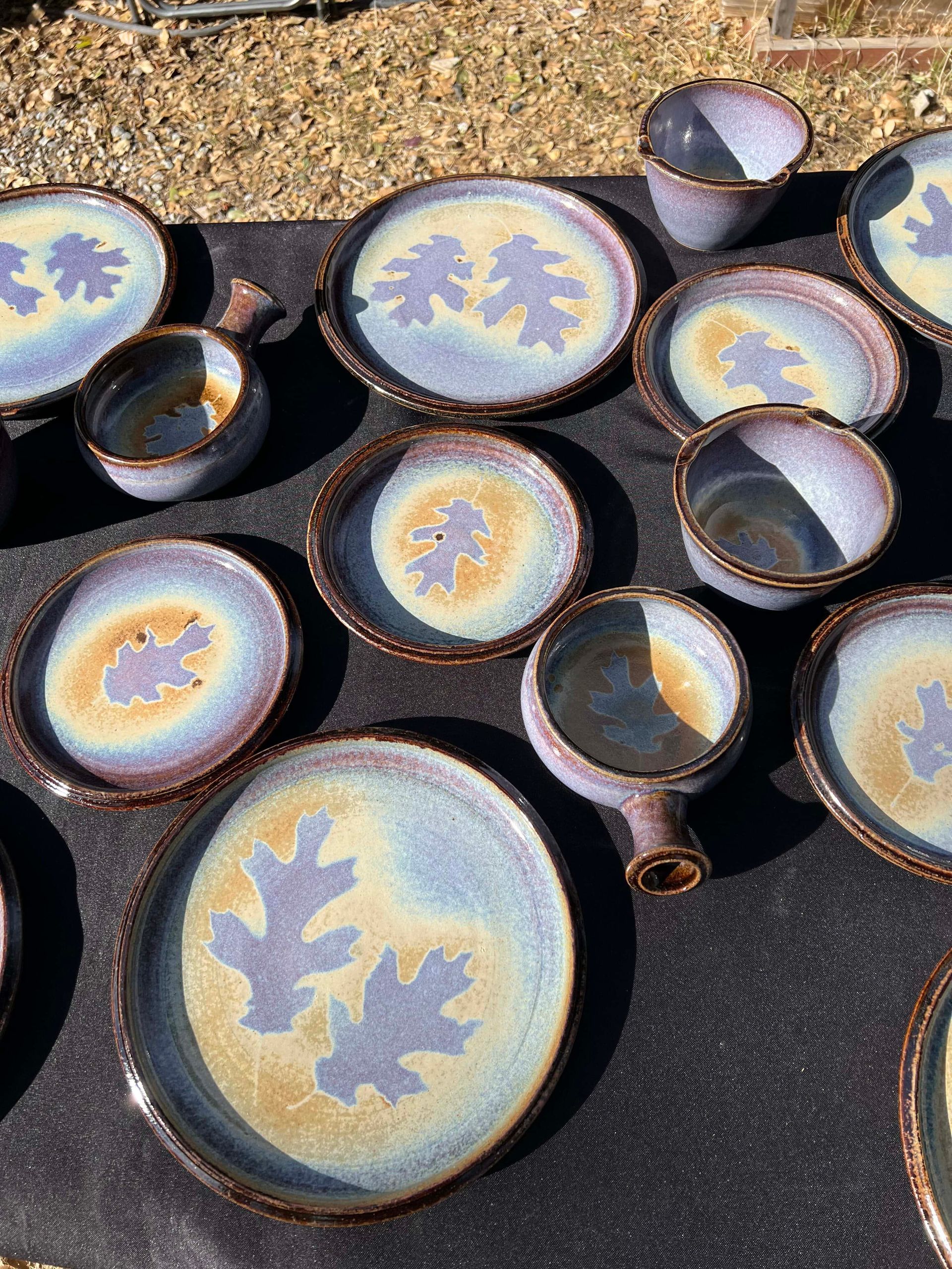 Set of plates, bowls, and mugs in the colors purple, blue, and yellow, with the outline of an oak leaf in the center of each.
