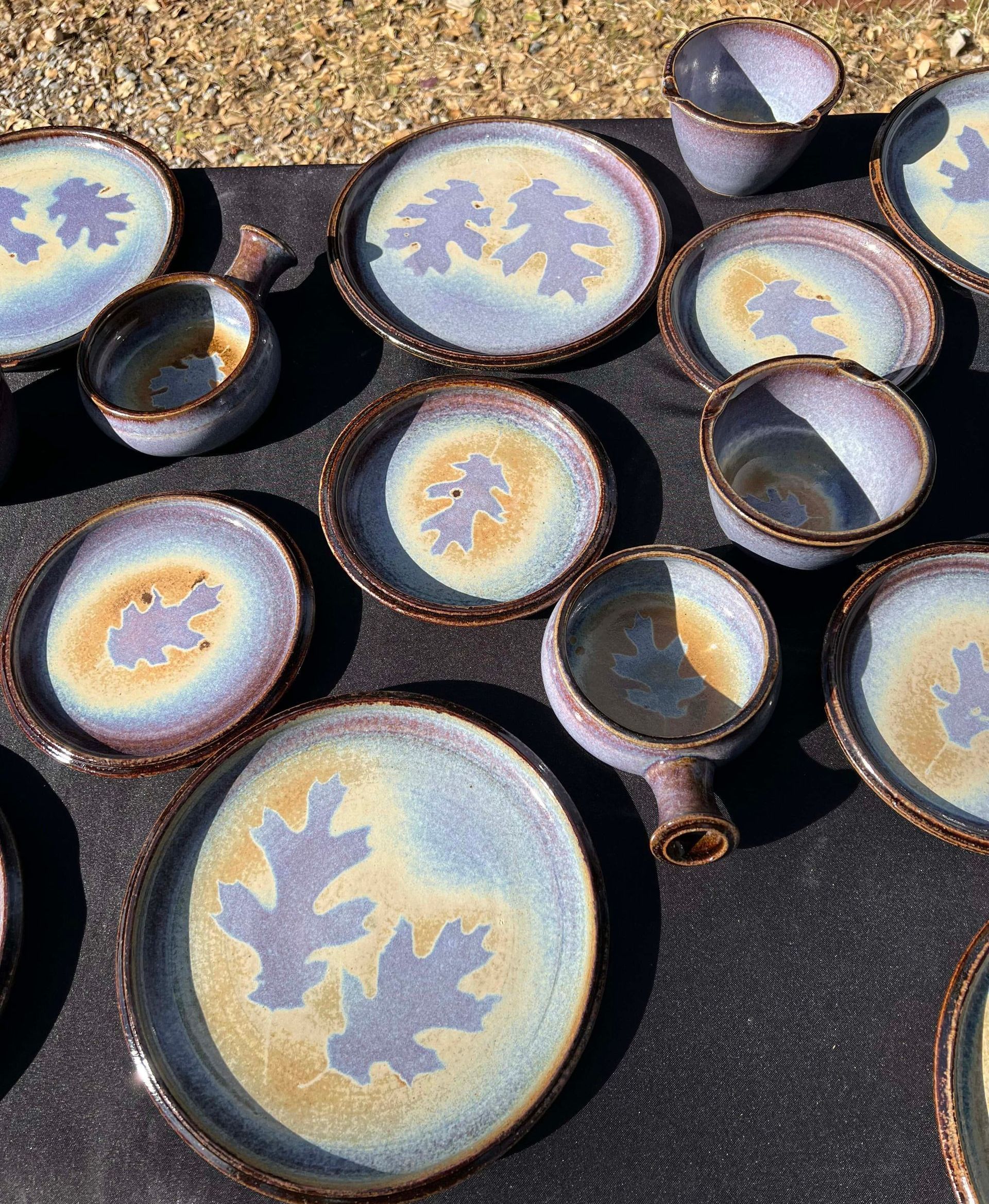 Set of plates, bowls, and mugs in the colors purple, blue, and yellow, with the outline of an oak leaf in the center of each.