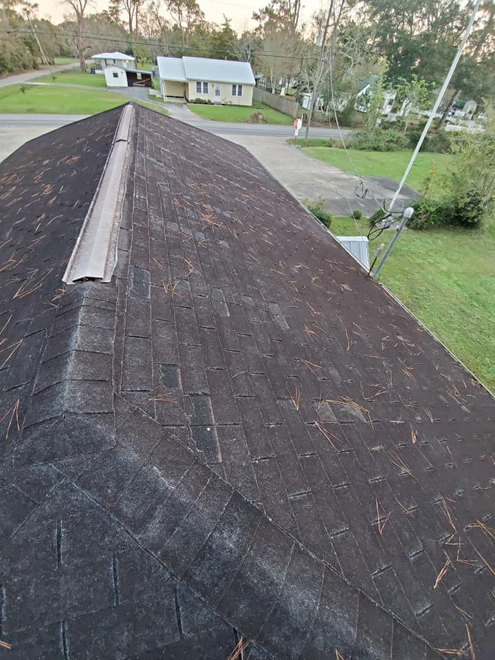 View of a dark, weathered shingled roof with a metal ridge cap, trees, and houses in the background.