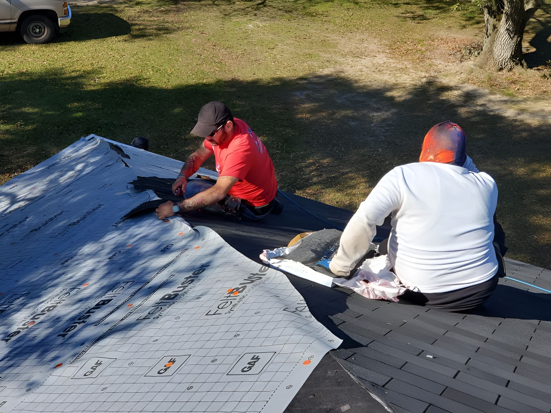 Two roofers in casual attire work on a roof; removing old shingles. Green grass and a tree in the background.