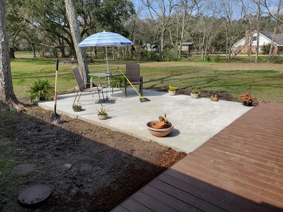 A concrete patio with a table, chairs, and umbrella in a yard.  Gardening tools and plants are nearby.