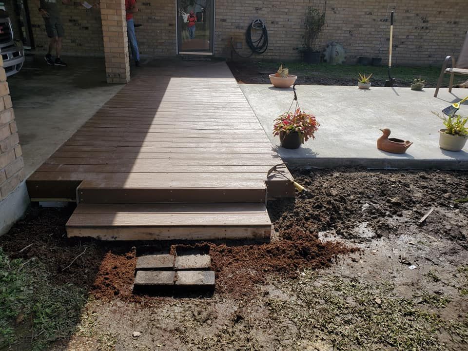 A brown wooden ramp and steps lead to a door from a patio. Ground with dirt and plants.