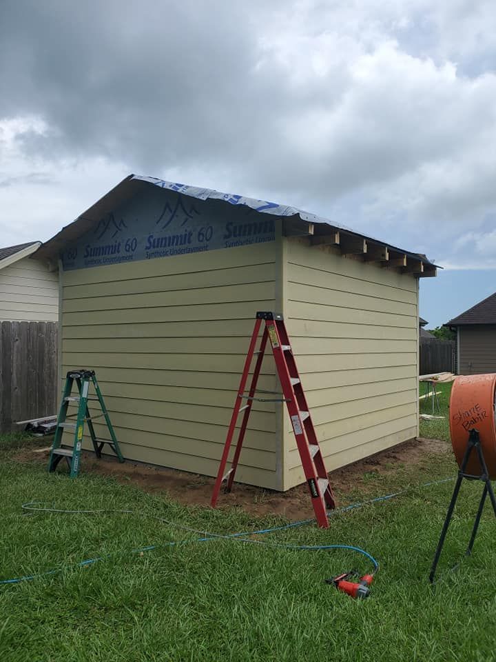 A tan shed under construction in a grassy yard, with a blue tarp on the roof and ladders leaning against the walls.