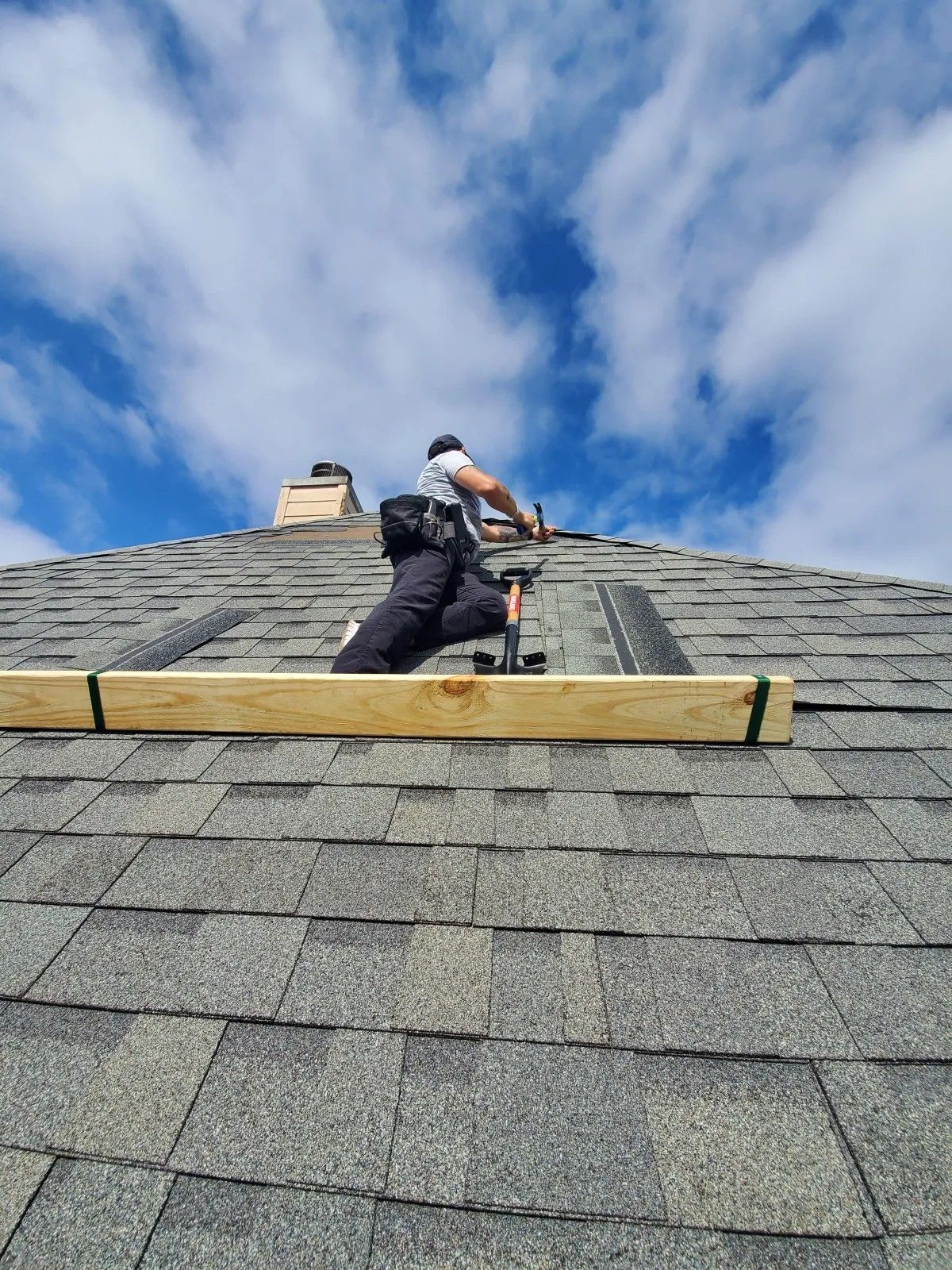 Roofer on a shingled roof under a partly cloudy sky, using tools. Wooden plank secured by green straps.