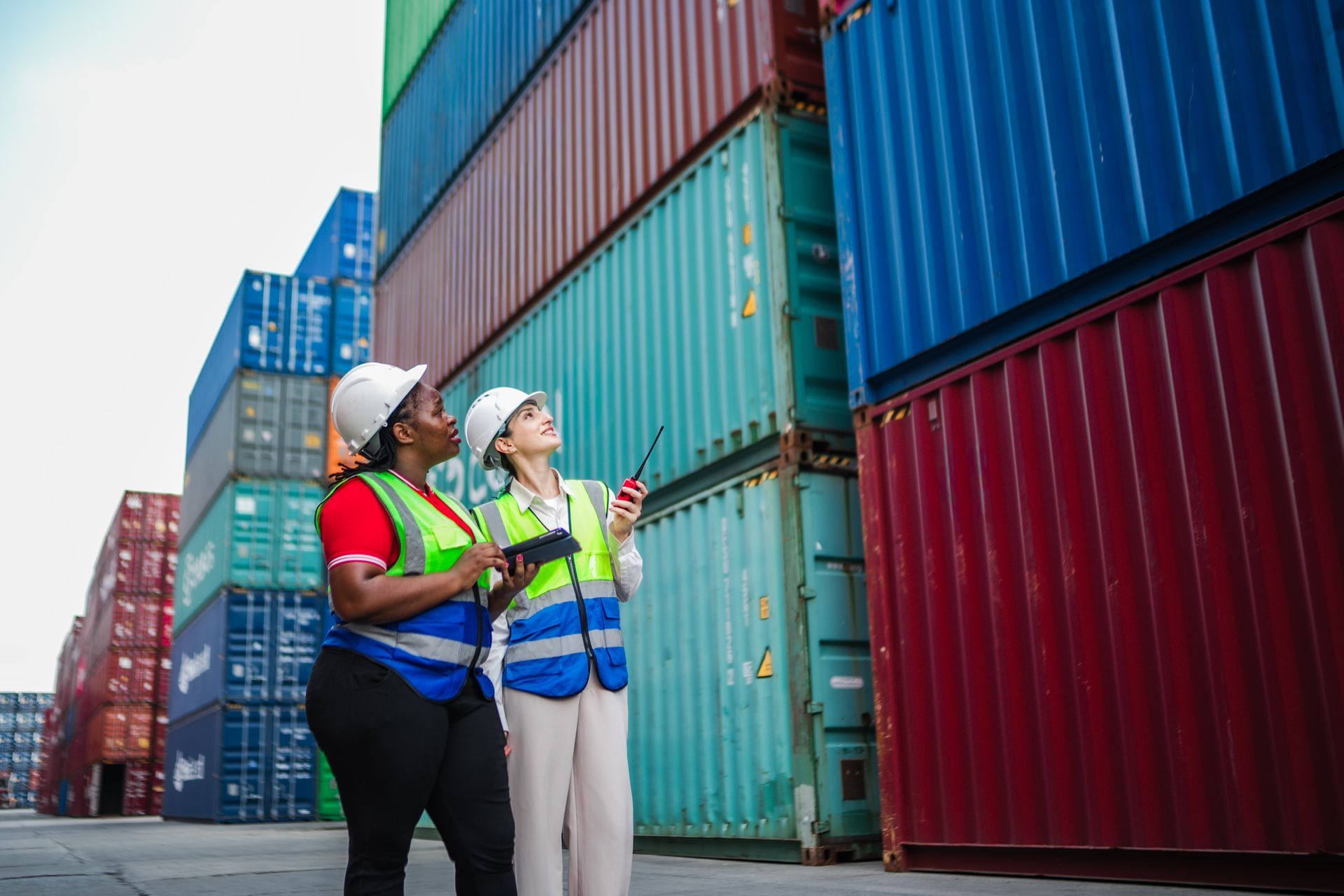 Workers inspect stacked shipping containers at a busy logistics yard.