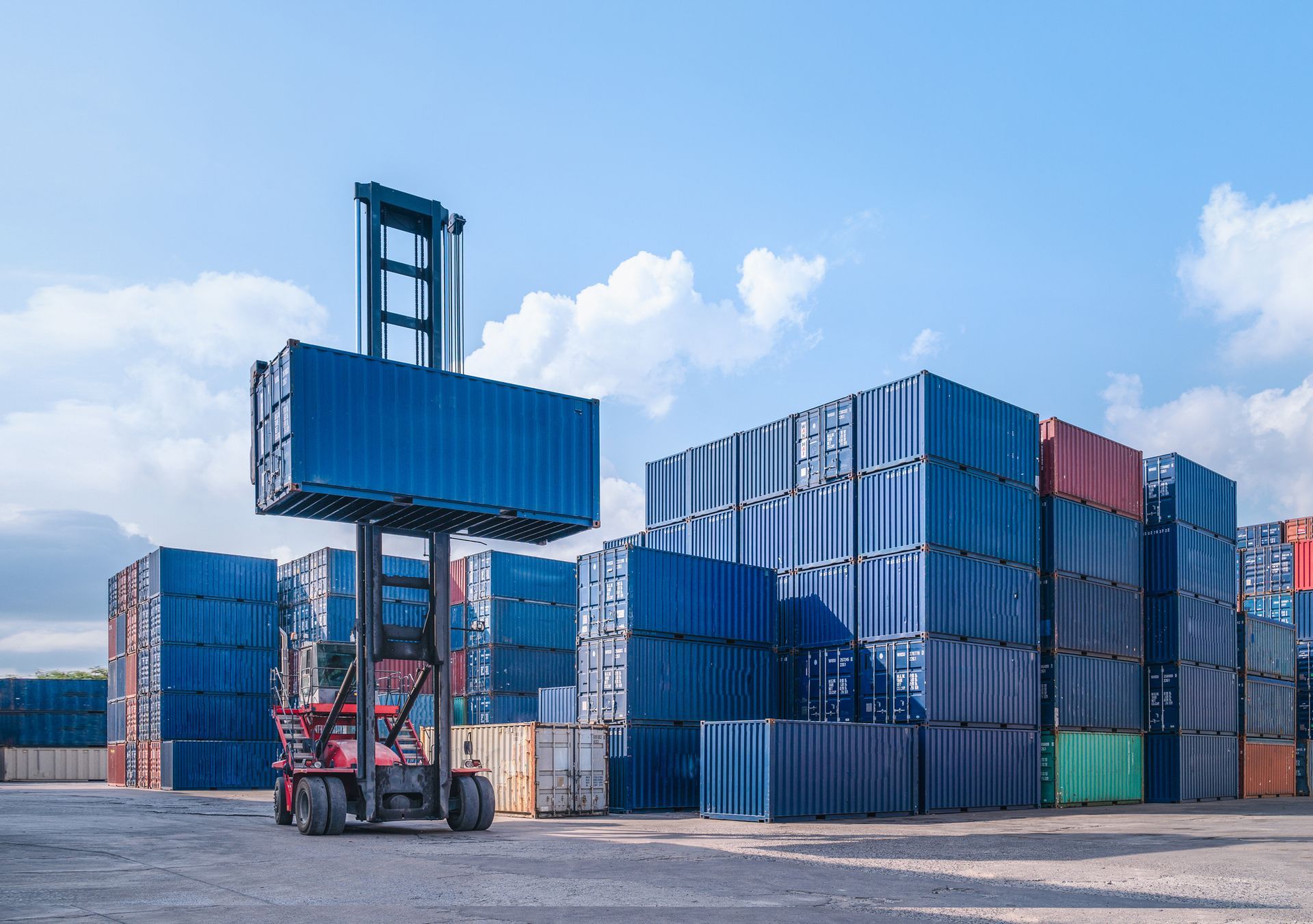 A forklift lifts a blue shipping container in a yard with stacked units.