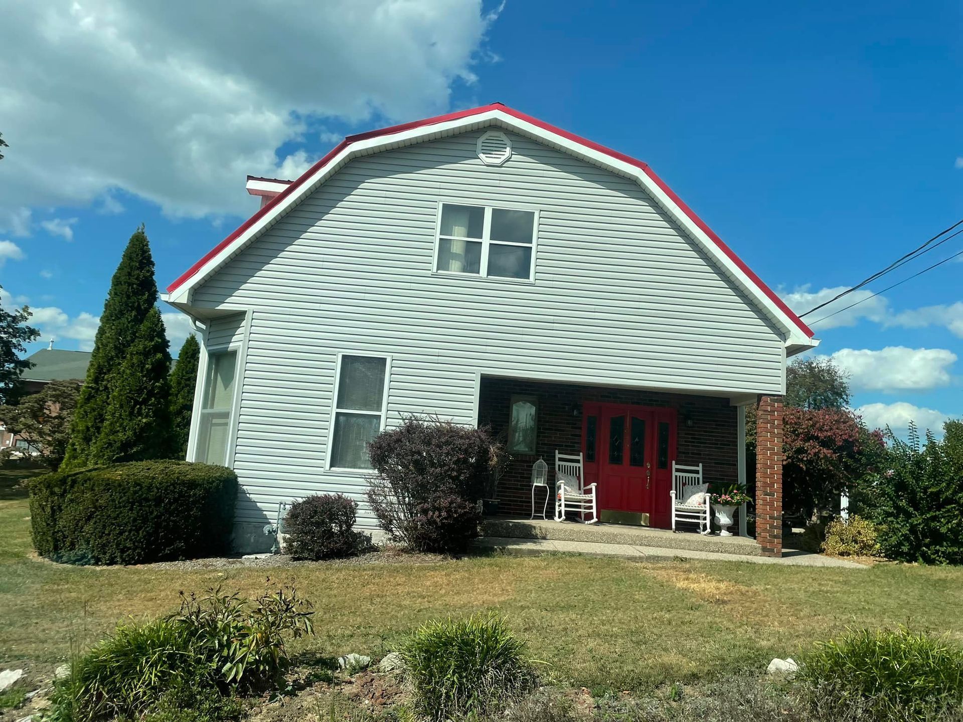 A two-story grey-sided house with a barn-style roof, a red door, a covered porch, and two rocking chairs.