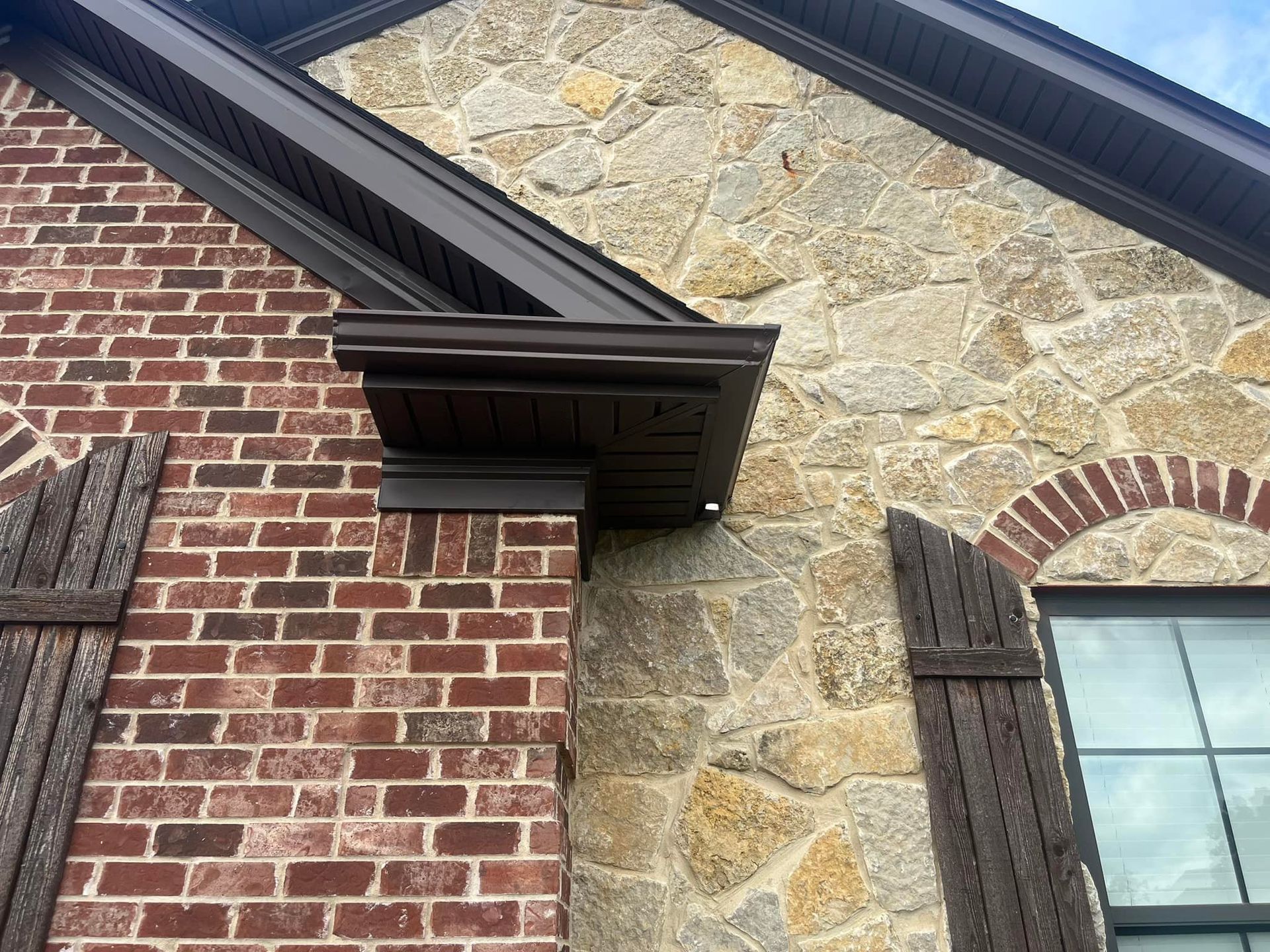 Exterior view of a home corner featuring red brick, natural stone cladding, dark brown shutters, and dark wood trim.