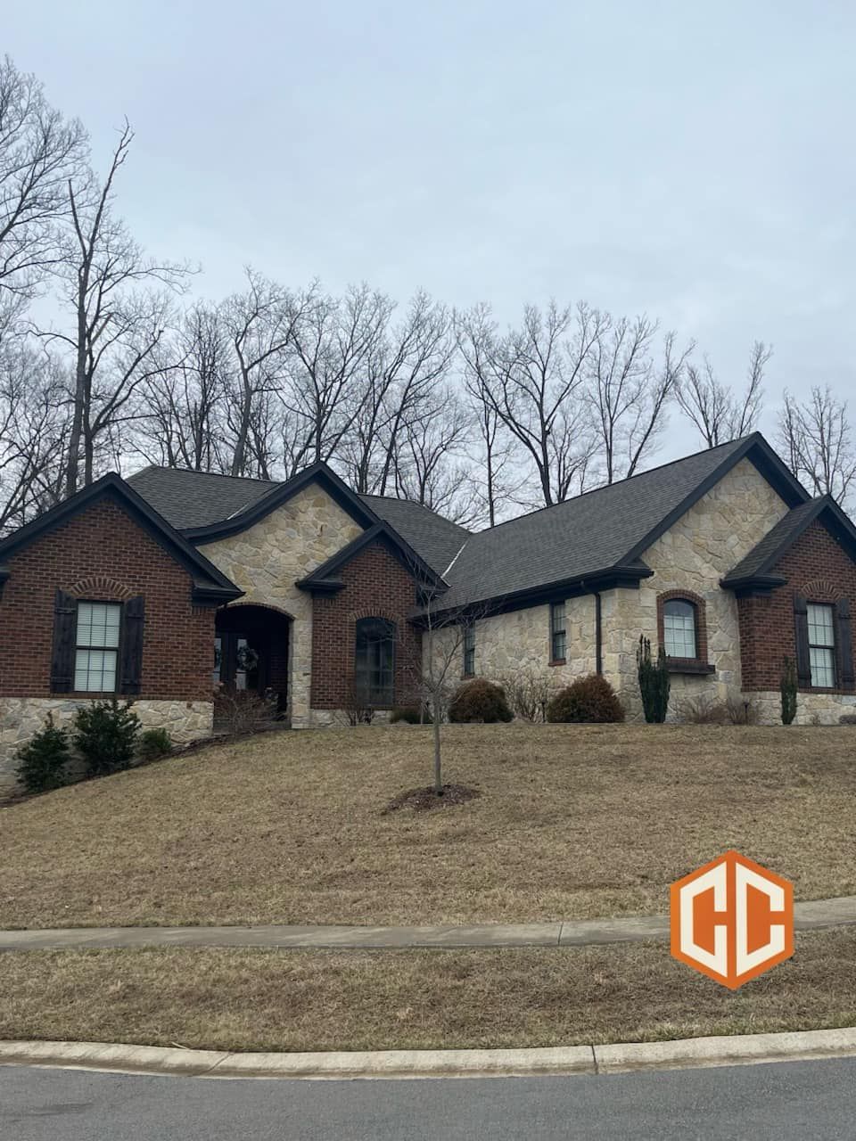 A one-story suburban home with a mix of light stone and dark brick siding under a gray roof, set on a sloped grassy lawn.