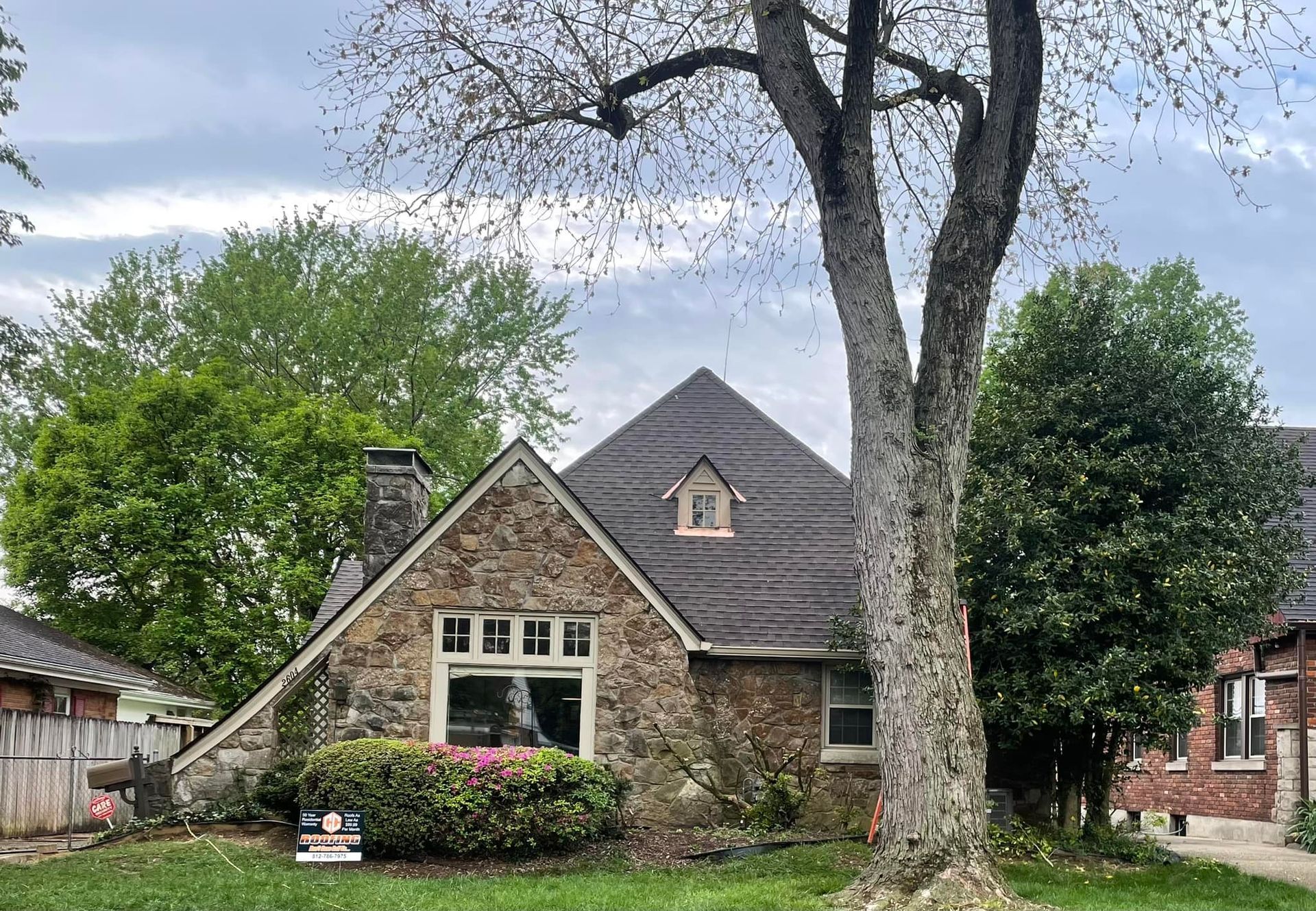 A stone-faced house with a steep, dark roof, featuring a dormer window, surrounded by trees and a green lawn.