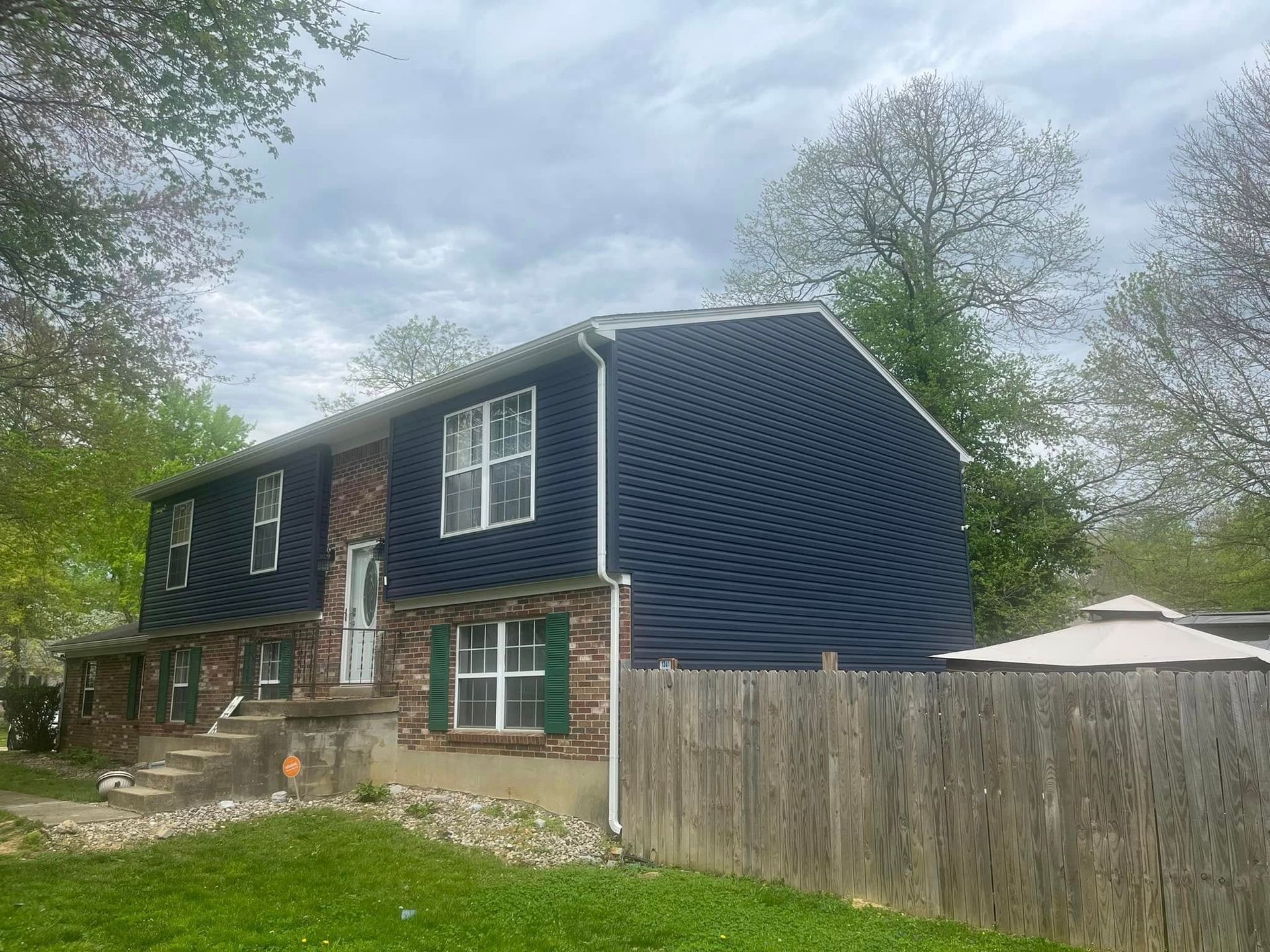 Two-story house with dark blue siding, brick accents, and green shutters, viewed from the yard on an overcast day.
