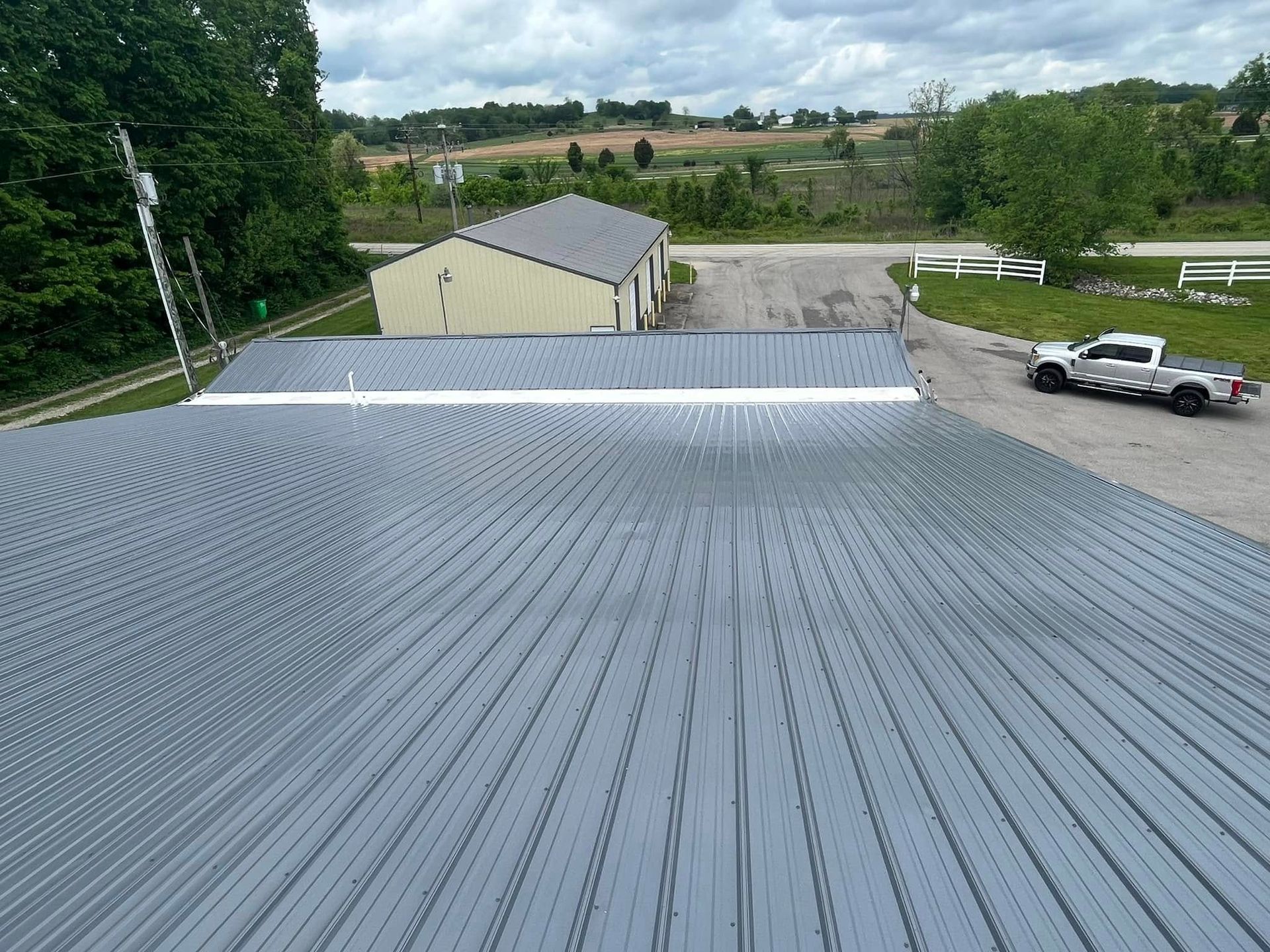 An elevated view of a gray metal roof on a sunny day, with a small building and a pickup truck visible in the background.