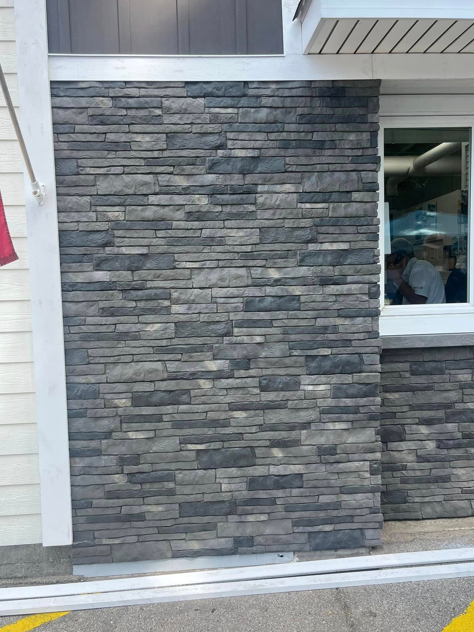 A wall facade featuring textured, dark gray stacked stone, situated next to a window and white siding.