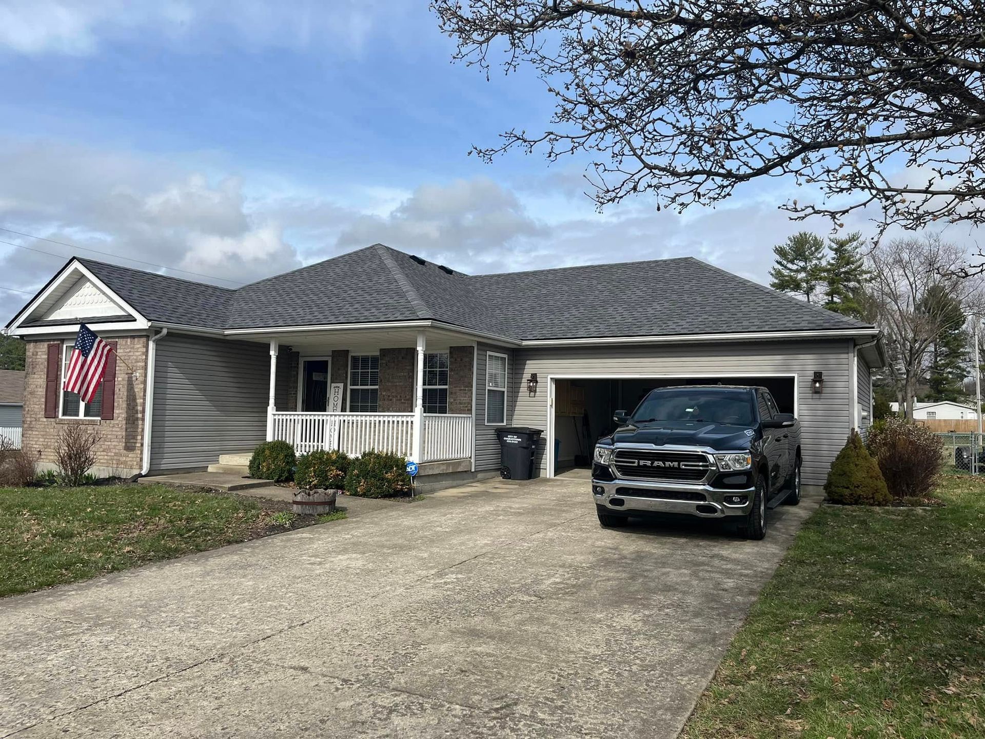 A gray, one-story house with a white front porch, brick accents, and an American flag, with a black truck in the garage.