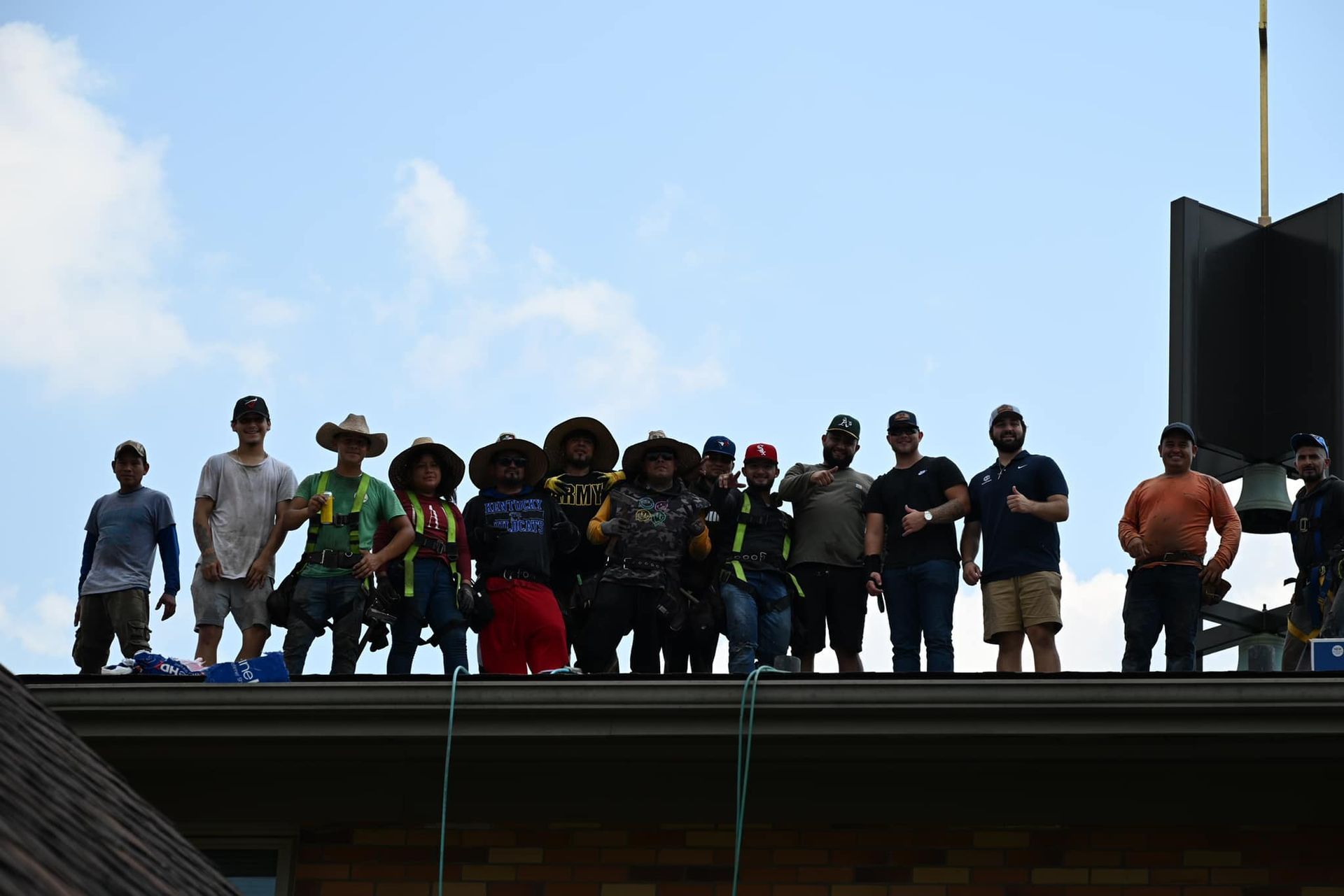 A group of people standing in a row on a rooftop against a clear blue sky, many wearing hats and work gear.