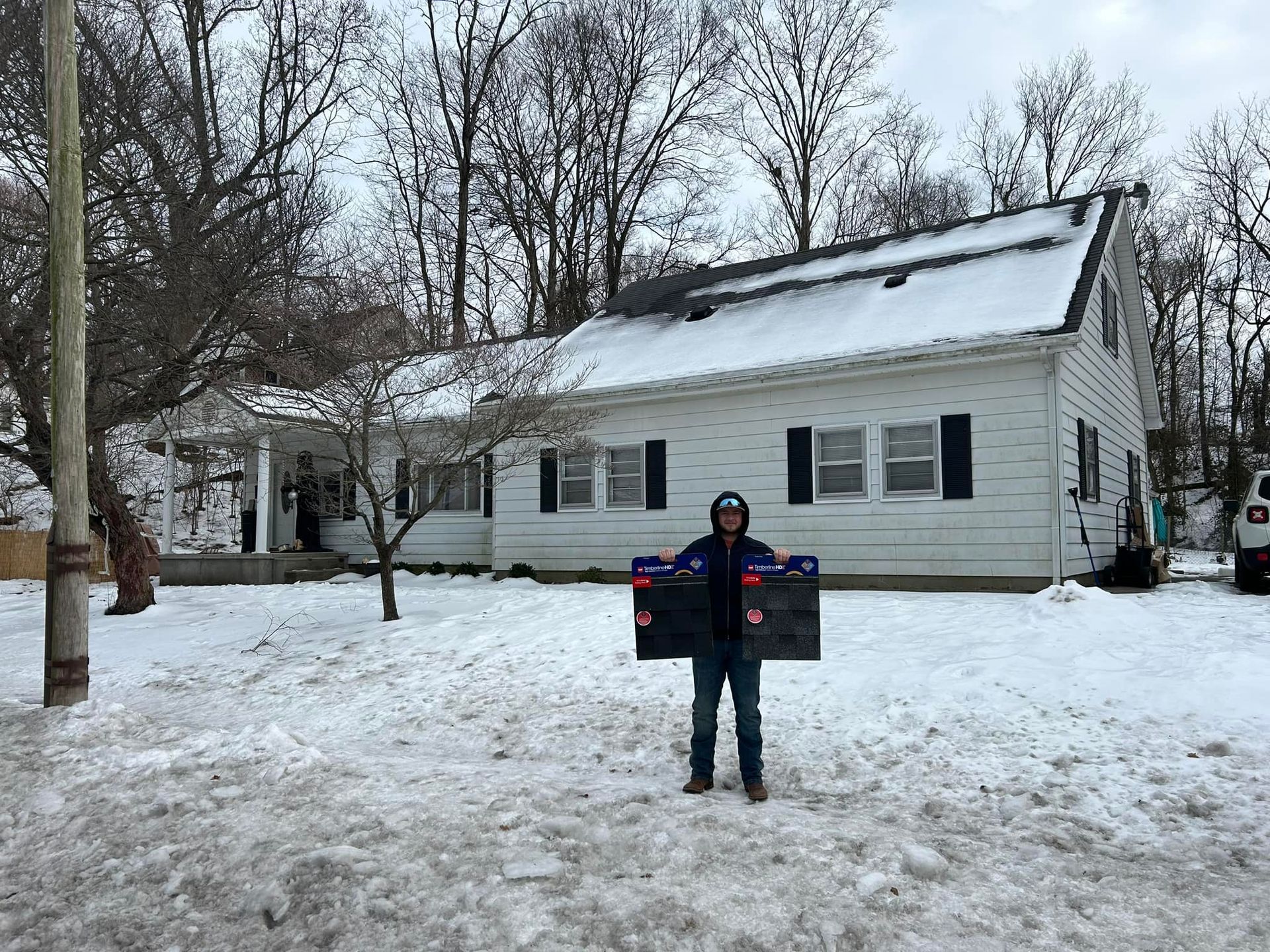 A person stands in a snowy yard in front of a white house, holding two black panels in their outstretched hands.