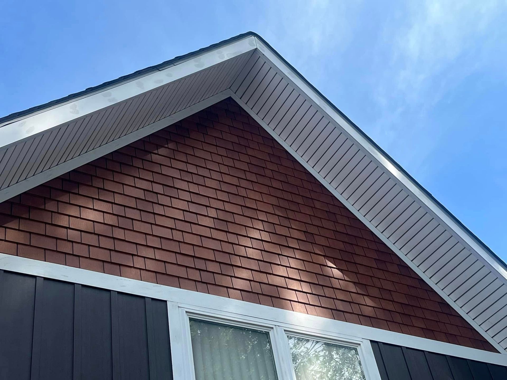 The pointed, shingled gable of a house with red siding, white trim, and a blue sky above.