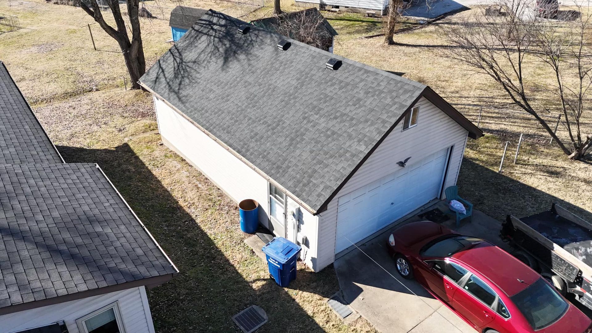 An aerial view of a detached garage with a dark roof and white siding, with a red car parked in the driveway nearby.