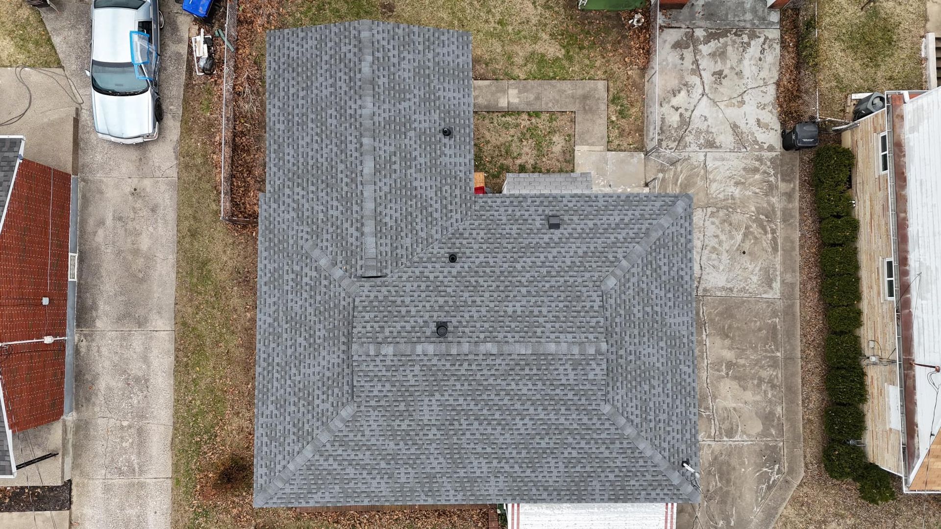 An aerial view of a gray shingled, L-shaped house roof in a residential neighborhood with surrounding concrete driveways.
