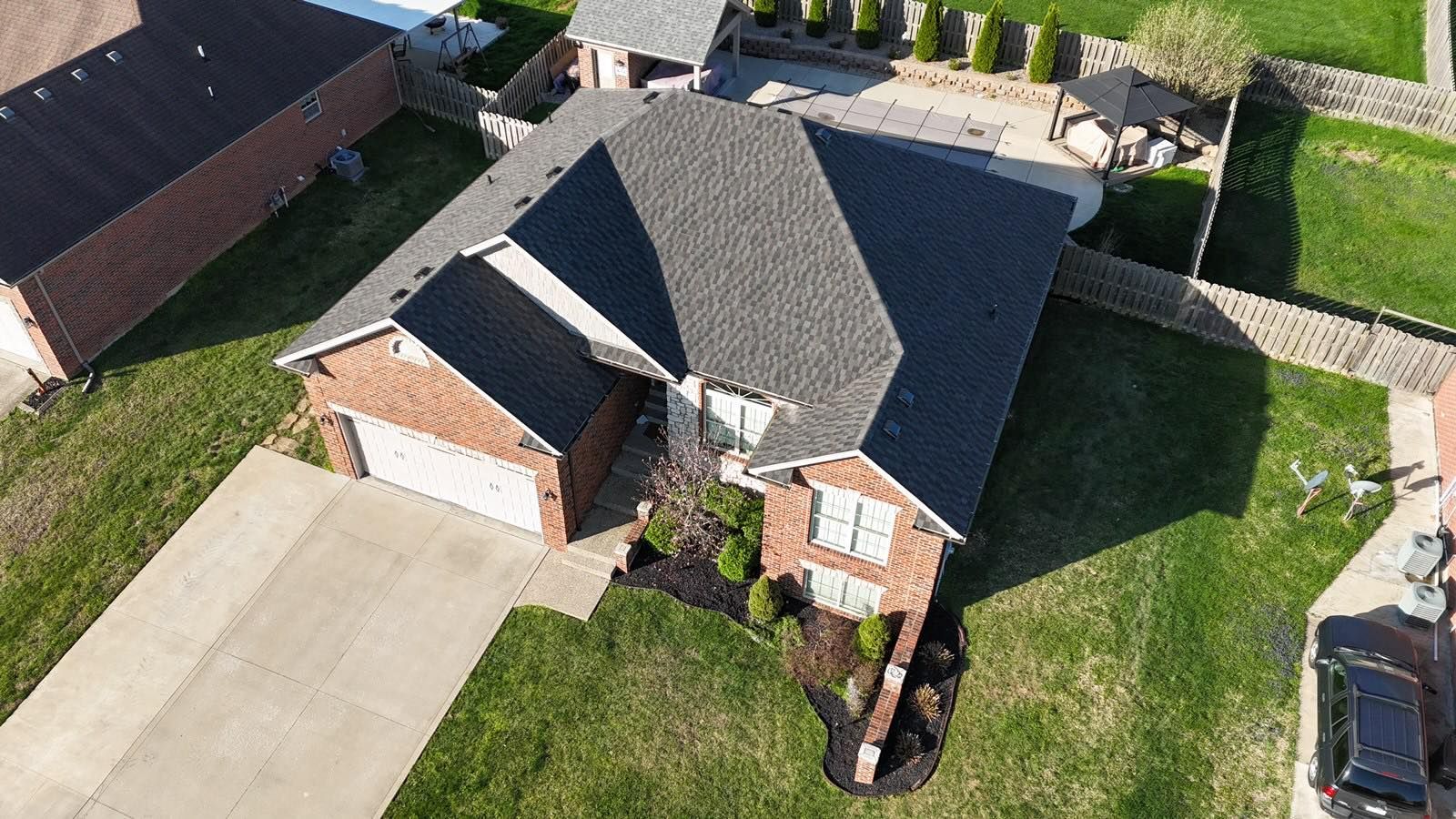 An aerial view of a brick house with a dark shingled roof, a paved driveway, and a backyard with a patio and pergola.