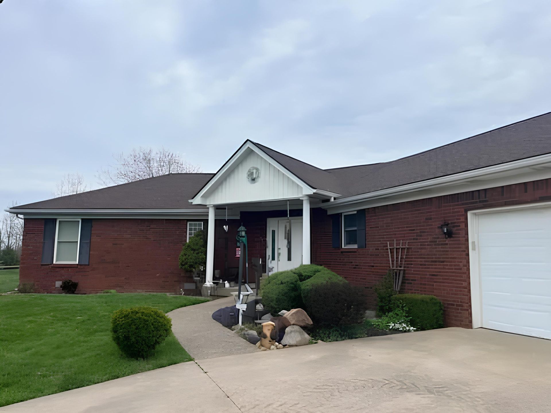 A single-story red brick house with a white gabled entryway, a paved walkway, and a white garage under a cloudy sky.