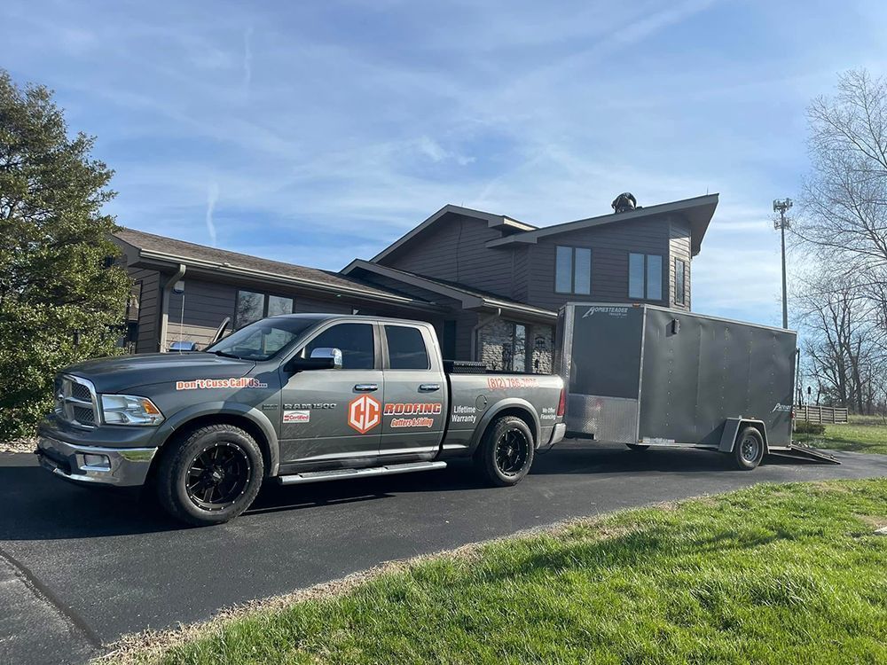 A grey pickup truck with a logo pulling a utility trailer parked on an asphalt driveway in front of a house.