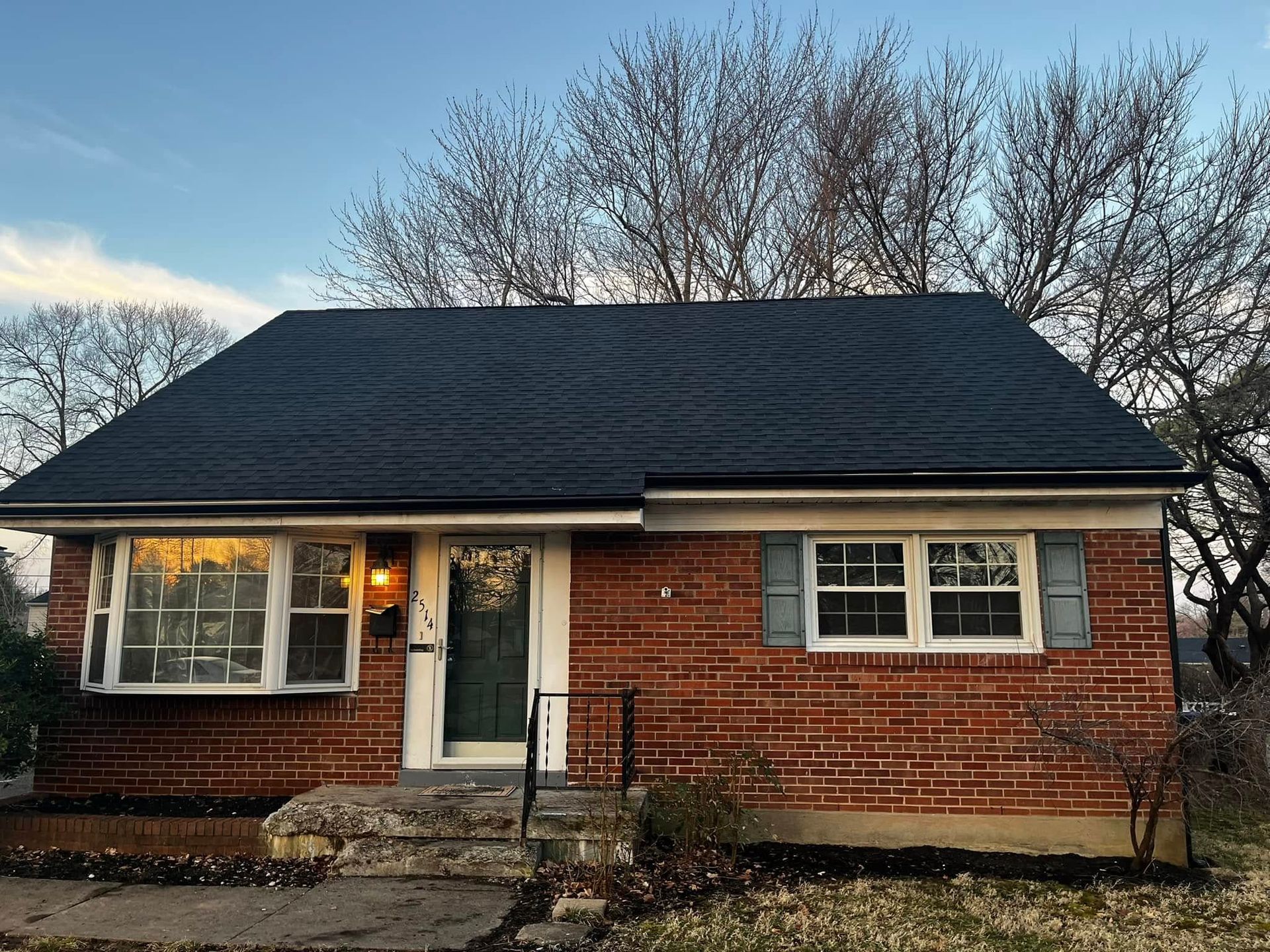 A single-story brick house with a dark gabled roof, a large bay window, and a green front door under a clear sky.