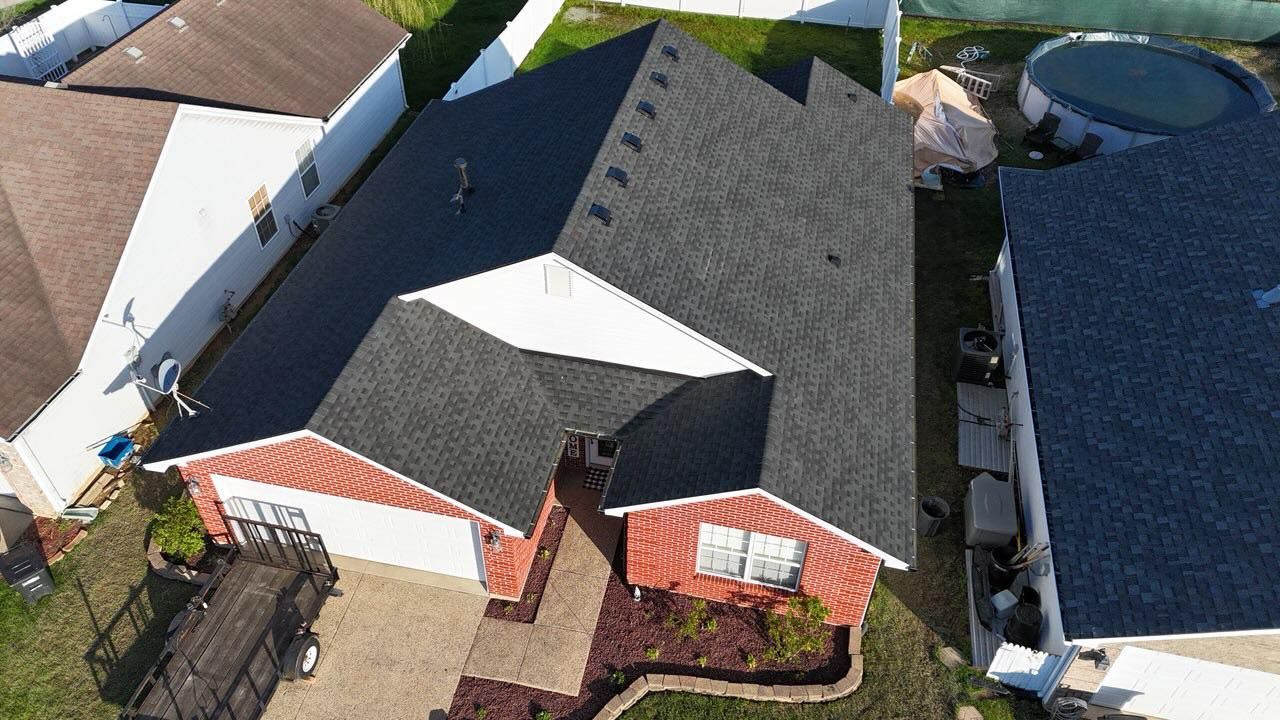 An aerial view of a brick house with a dark shingled roof, surrounded by neighboring homes and a backyard pool.