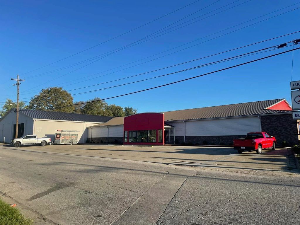 A commercial building with white walls, a dark roof, and a red front entrance, with two trucks parked in the lot.