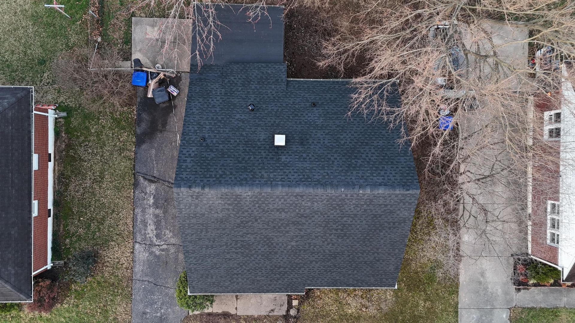 An aerial view of a dark-shingled house roof with a vent, surrounded by a lawn, paved driveway, and bare trees.